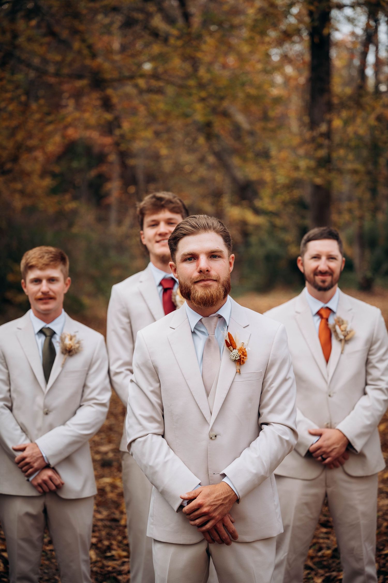 Wedding party in cream colored suits poses together against autumn foliage background.