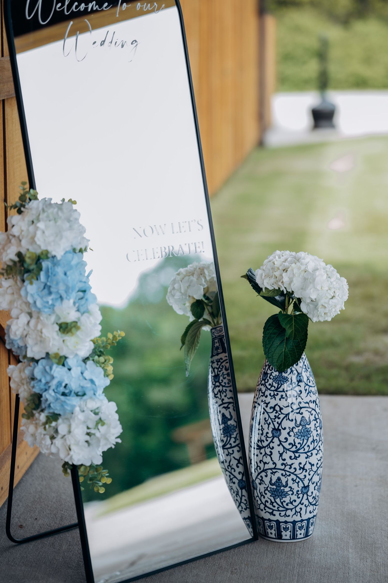 Wedding welcome mirror sign with blue hydrangeas beside a blue and white porcelain vase with white hydrangeas.