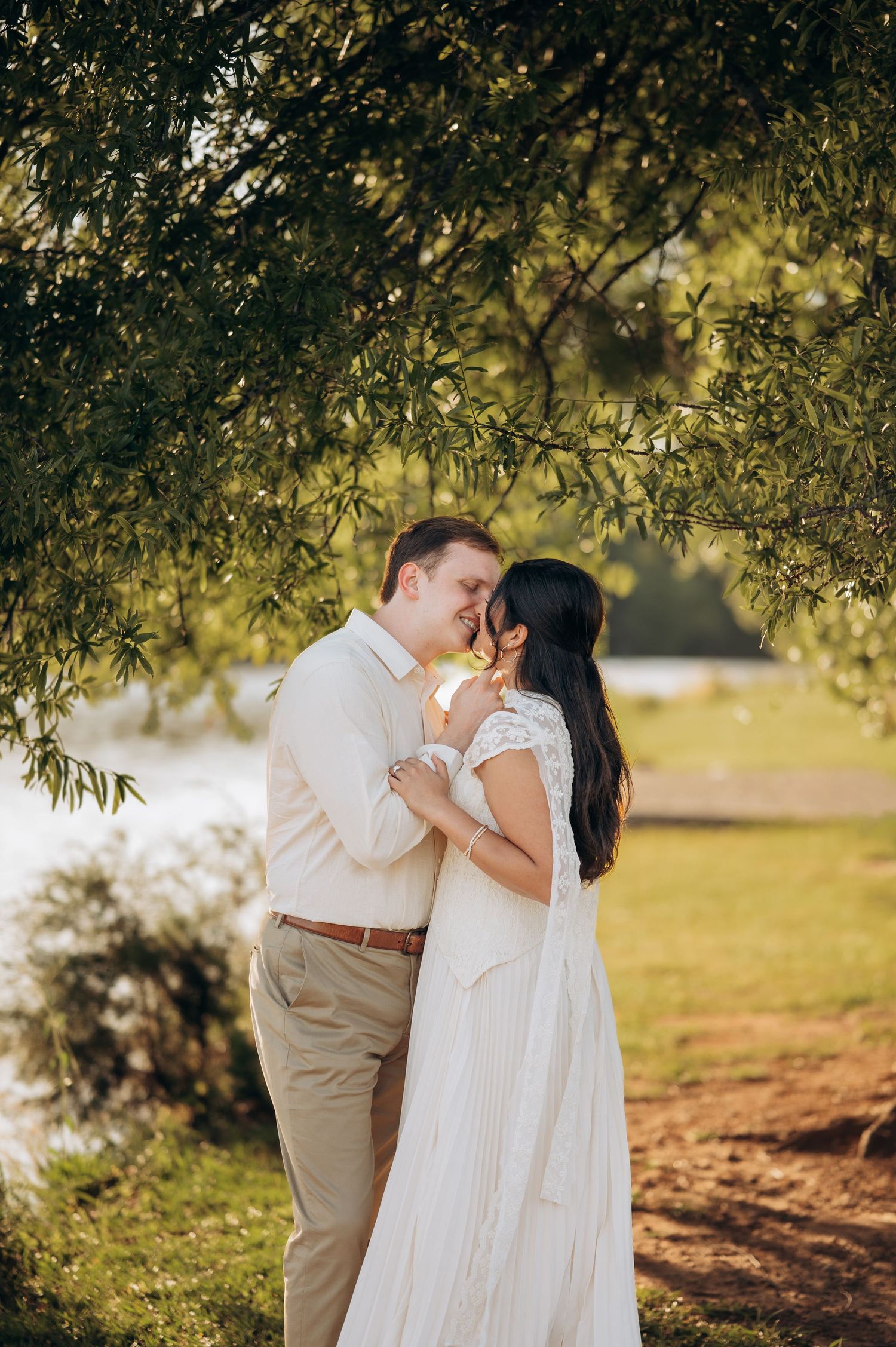 Couple sharing a romantic moment under a willow tree by a lake, bride in white lace dress, groom in khaki.