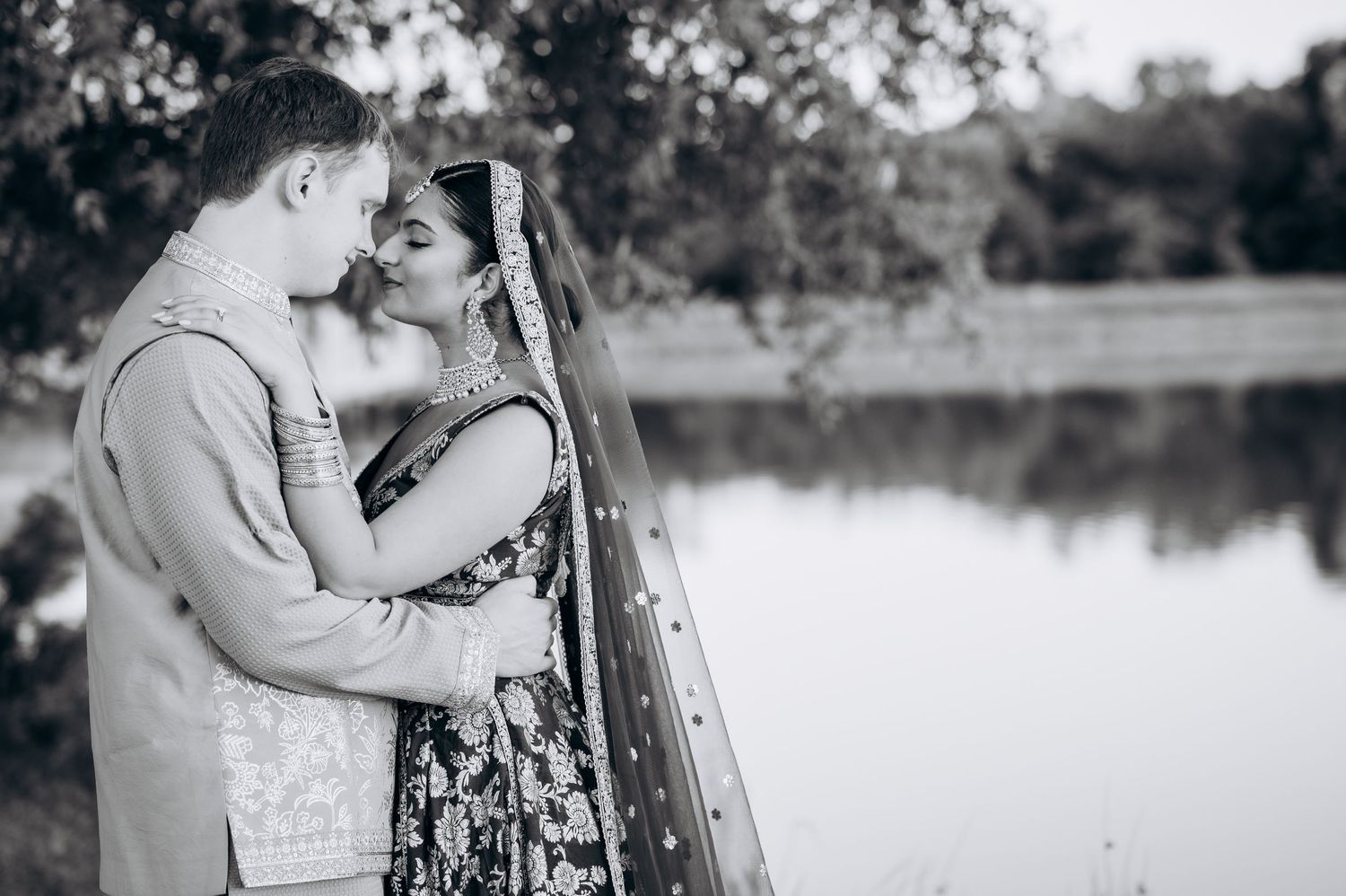 Black and white photo of a couple in traditional South Asian wedding attire sharing a romantic moment by a lake.
