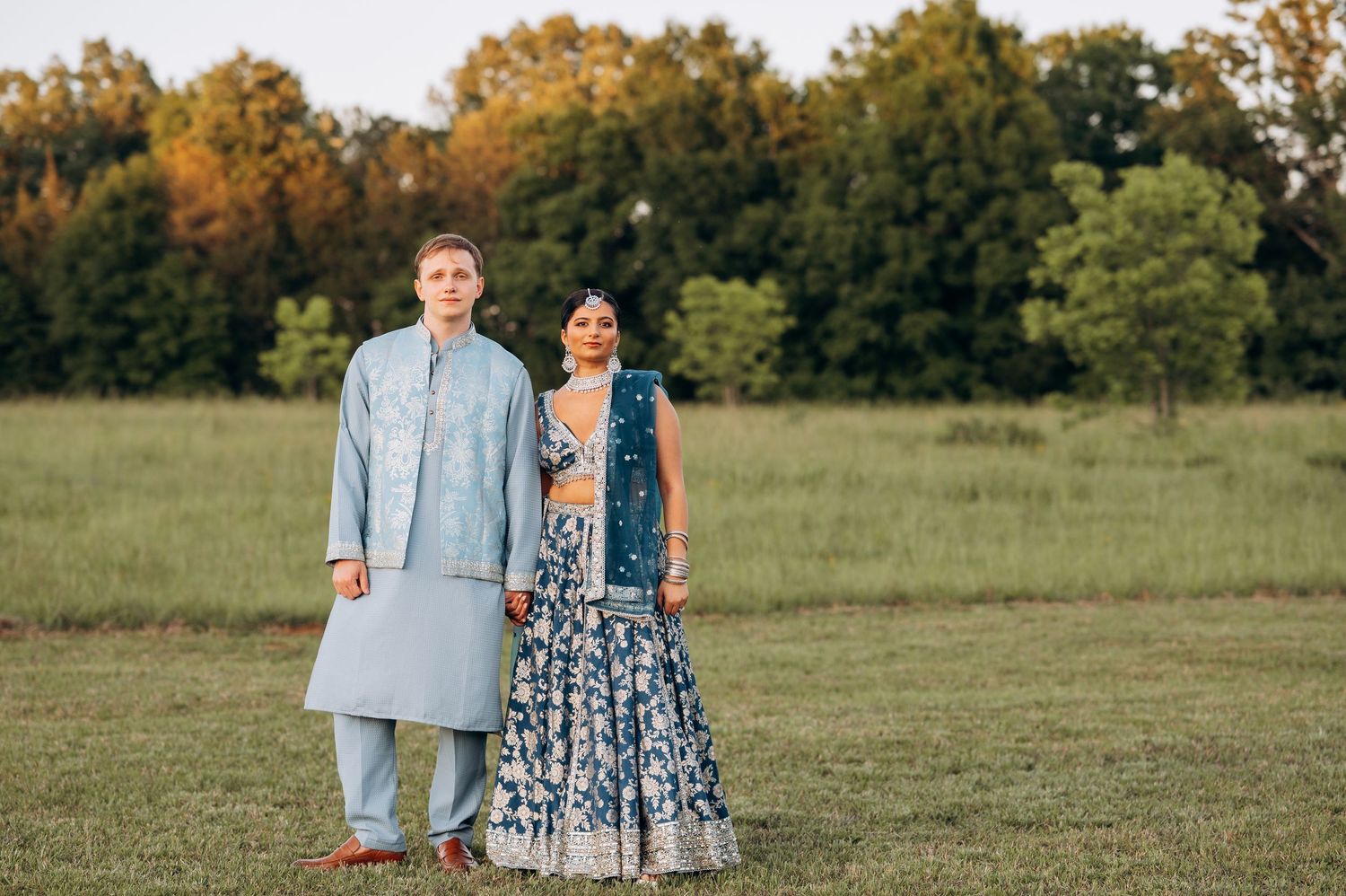 Couple in matching blue Indian wedding attire standing in a green field with trees in the background.