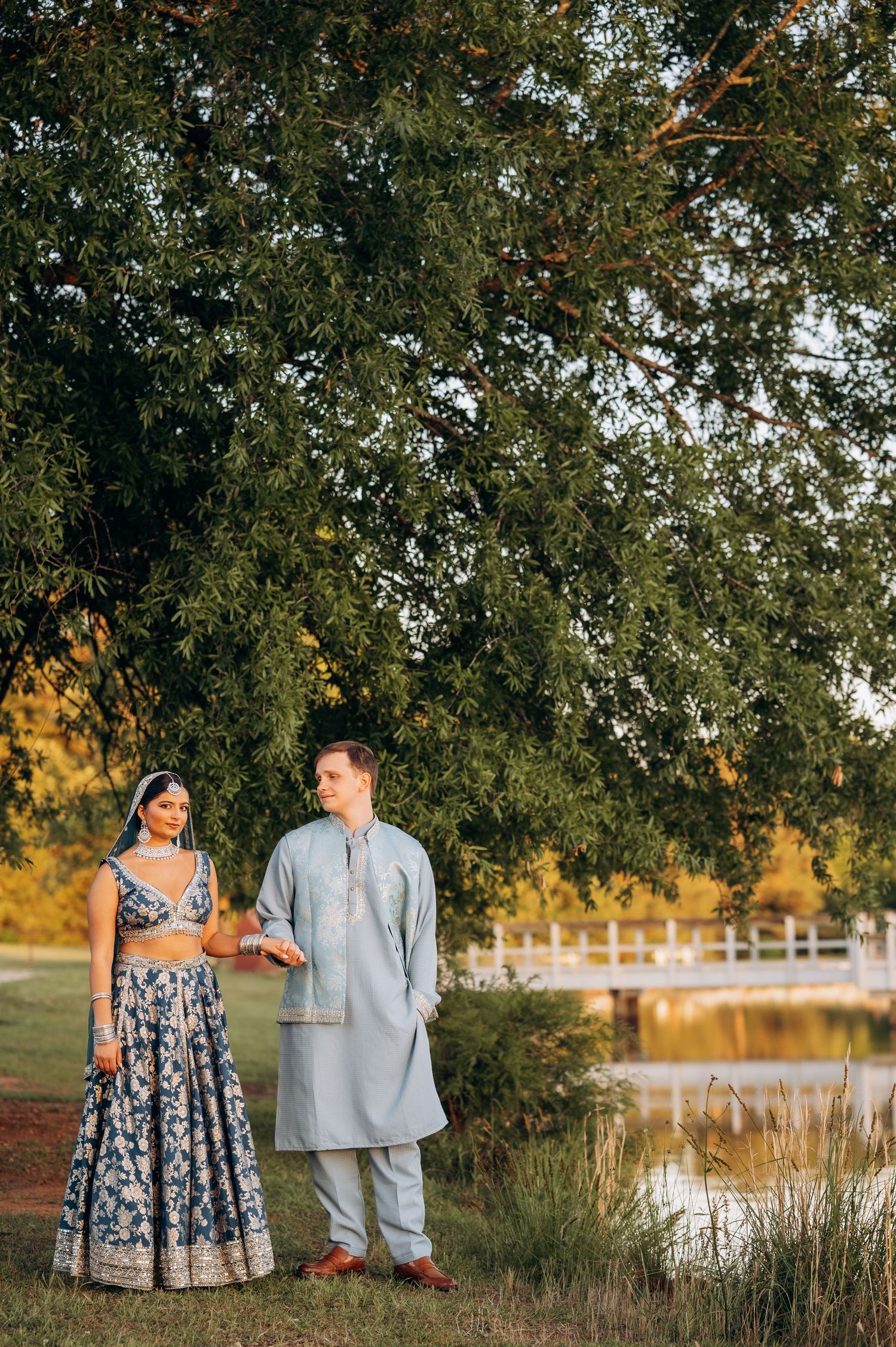Couple in traditional Indian wedding attire standing by a lakeside under a large tree at golden hour.