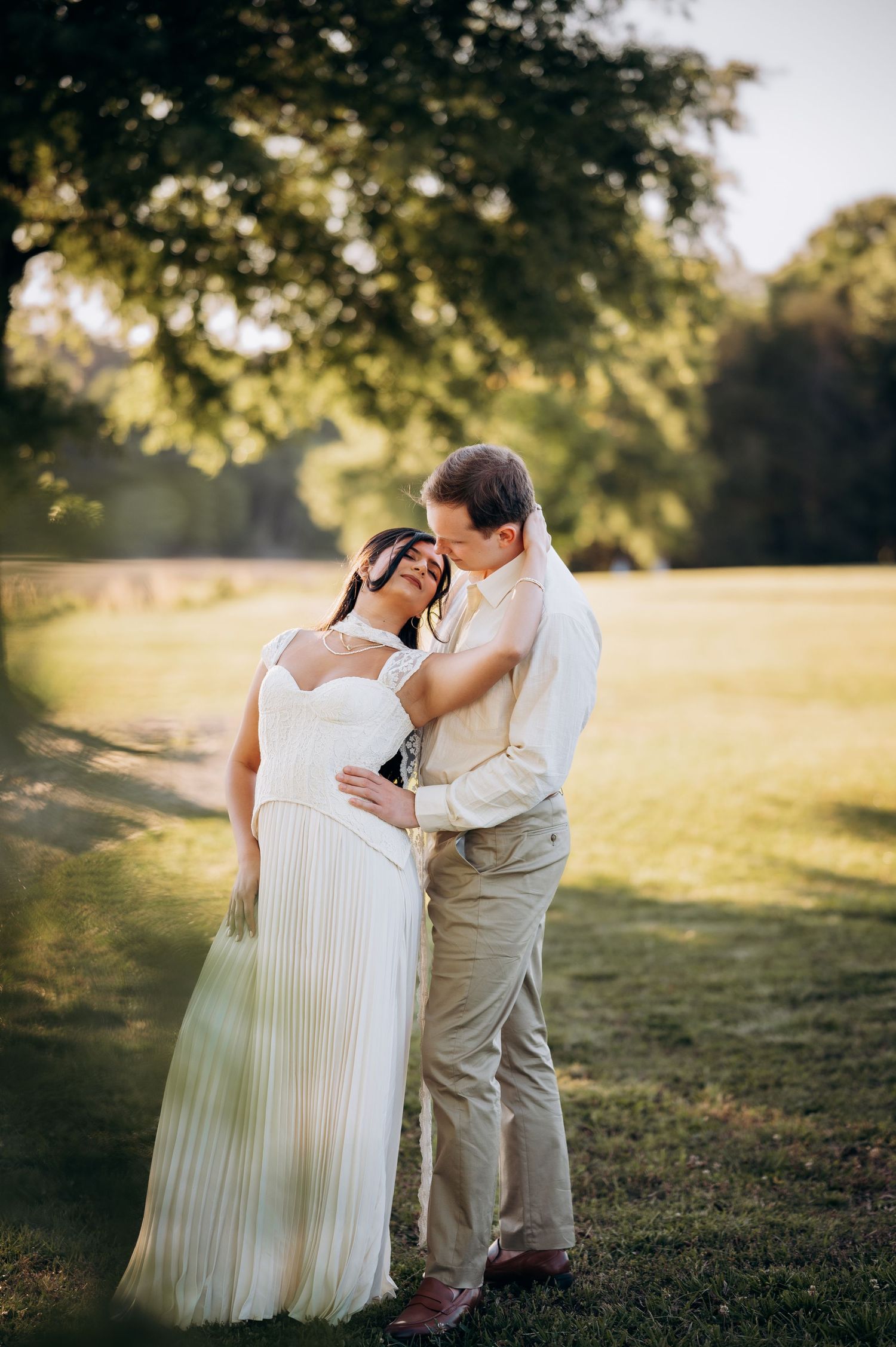 A couple shares a tender embrace in golden hour sunlight on a lush green lawn, bride in white gown, groom in tan suit.
