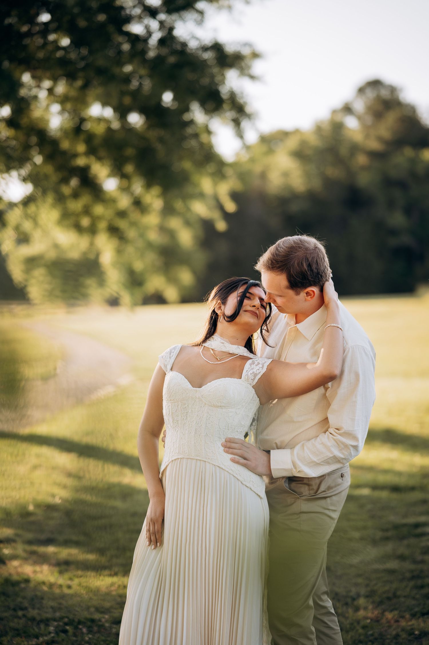 Couple embracing in golden sunlight outdoors, bride in white pleated gown, groom in cream shirt, lush green trees background.
