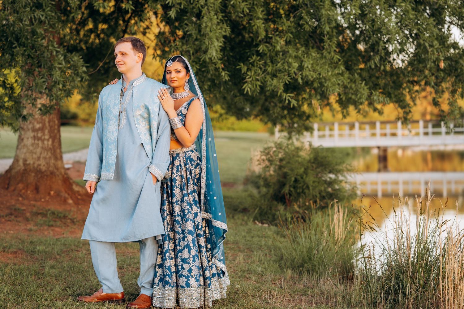 Couple in Indian wedding attire poses outdoors near a pond, man in light blue sherwani, woman in blue lehenga.