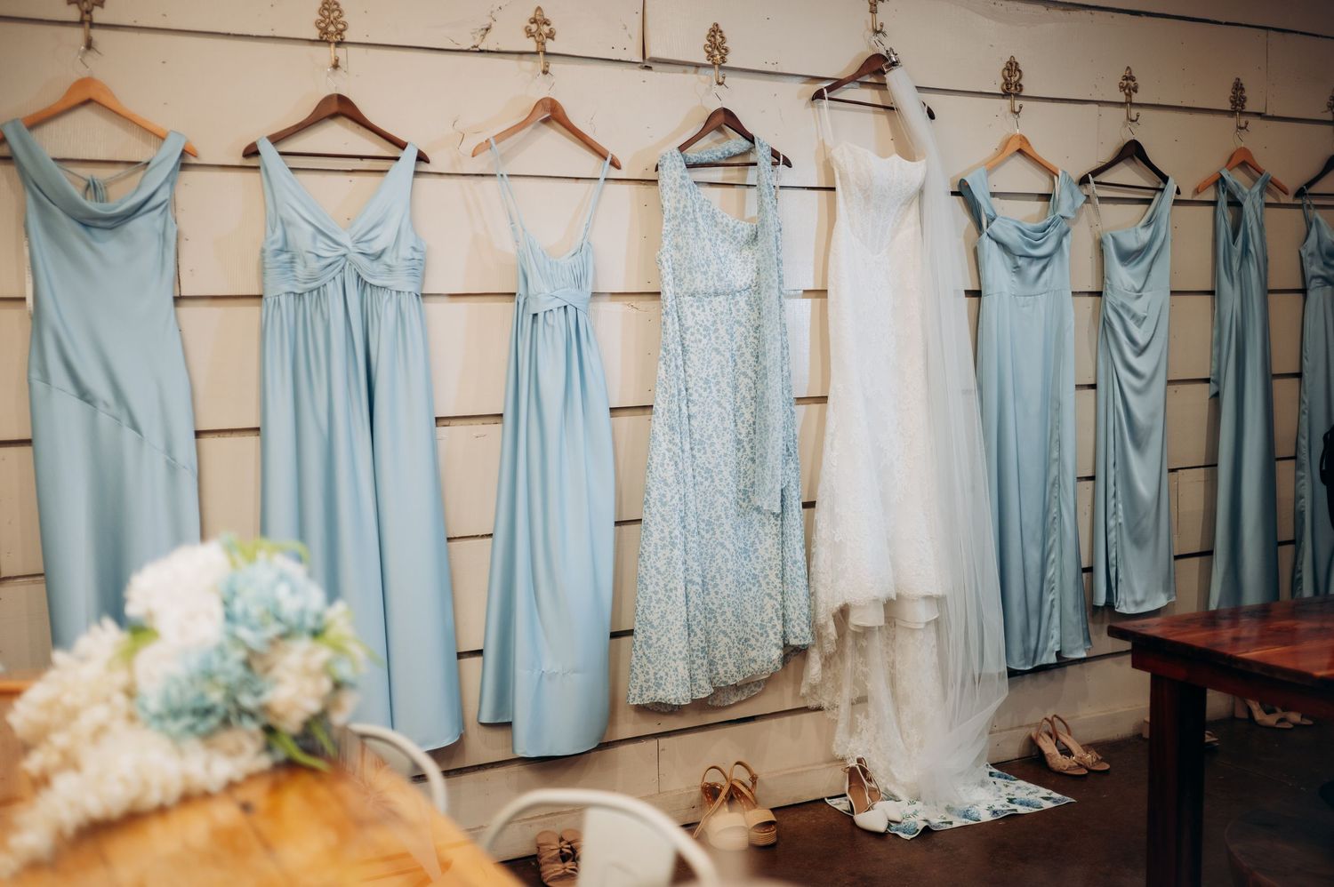 Light blue bridesmaid dresses and white bridal gown hanging on wooden wall hooks with bouquet and heels below.