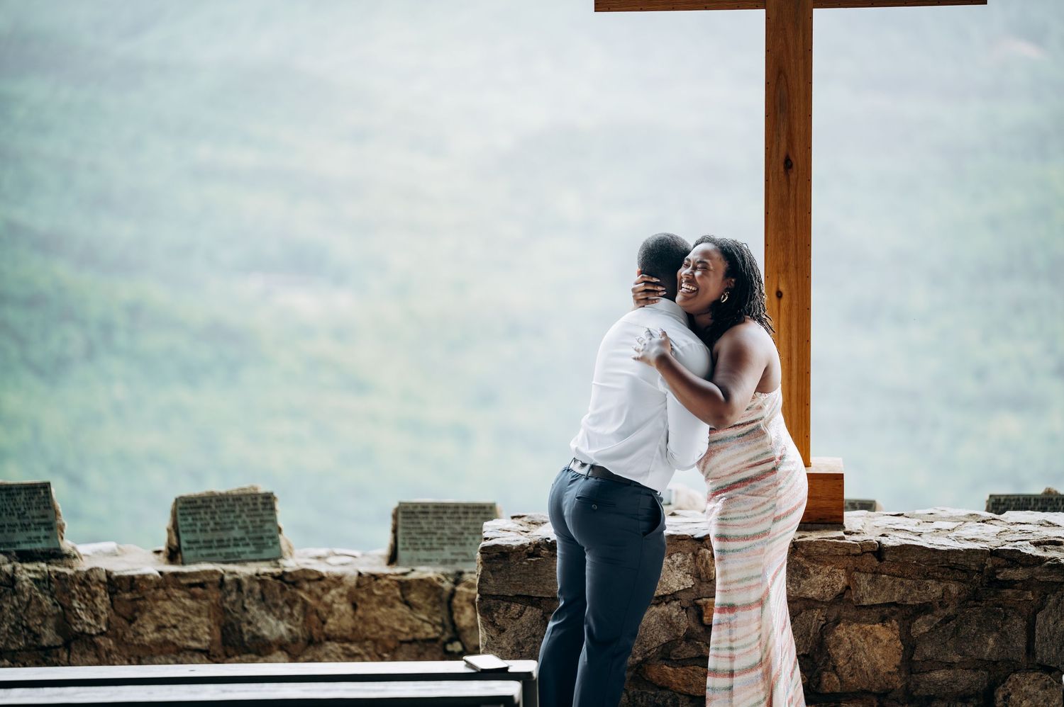 Couple embracing near a wooden cross on a stone terrace with lush green mountain views in the background.