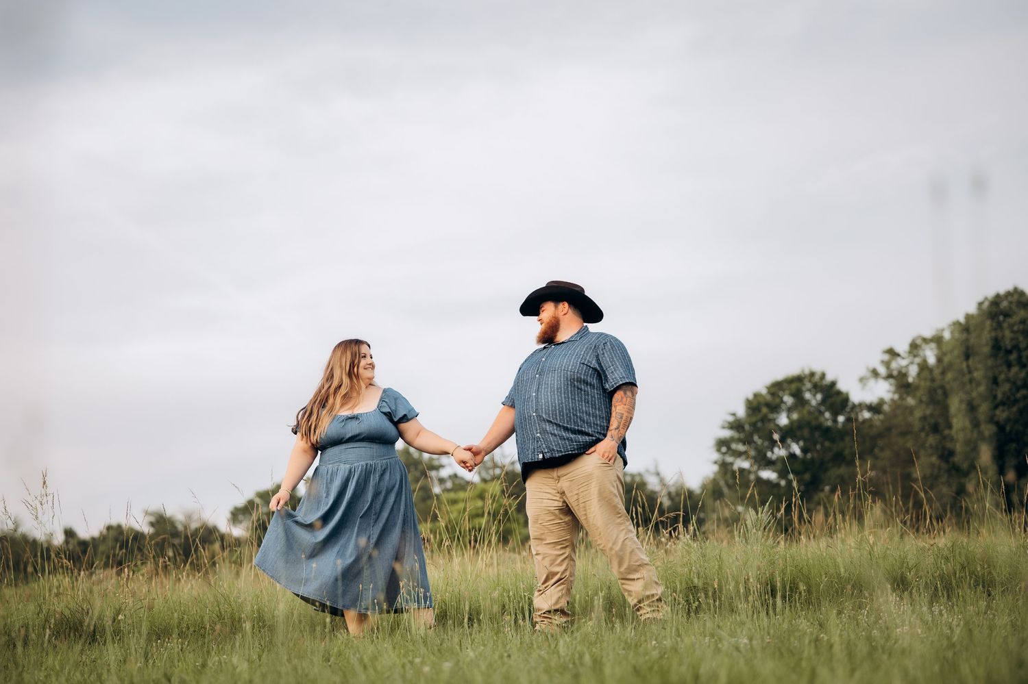 Couple holding hands walking through green field, woman in blue dress, man in cowboy hat and khakis, cloudy sky.