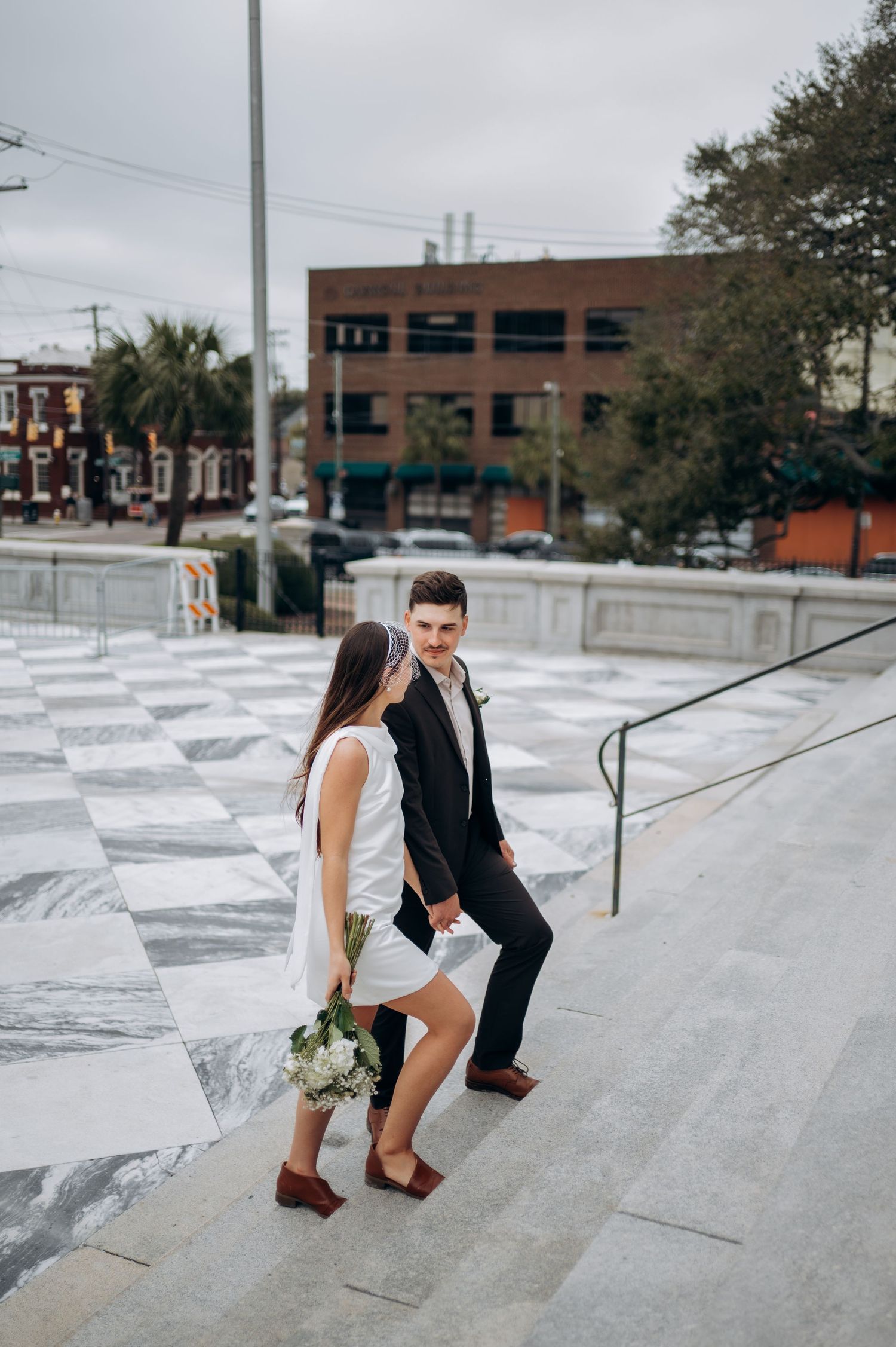 Newlywed couple holding hands walking up marble steps in an urban setting, bride carrying white bouquet.
