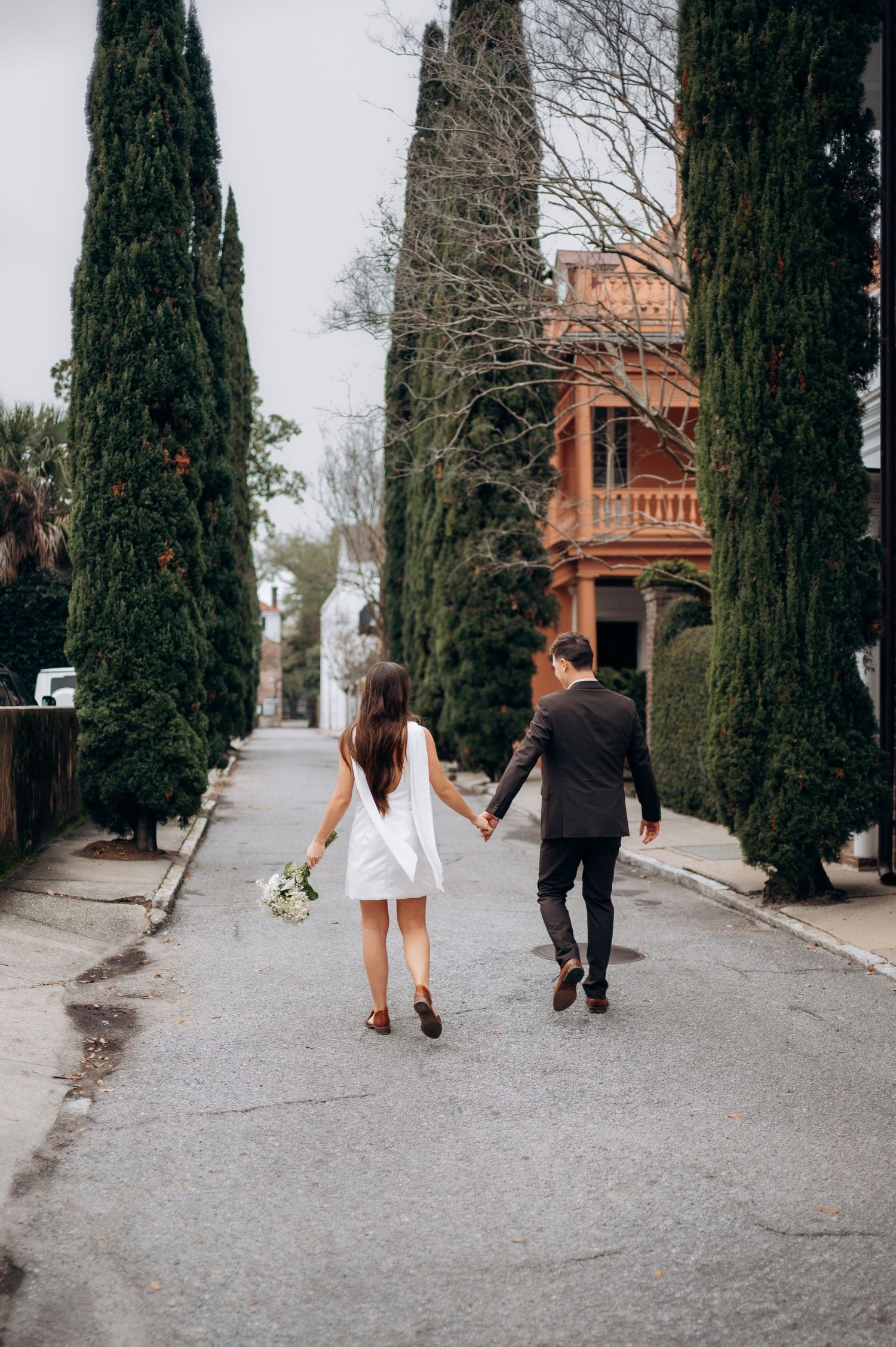 Bride and groom walking hand in hand down tree-lined street, bride carrying white bouquet, viewed from behind.