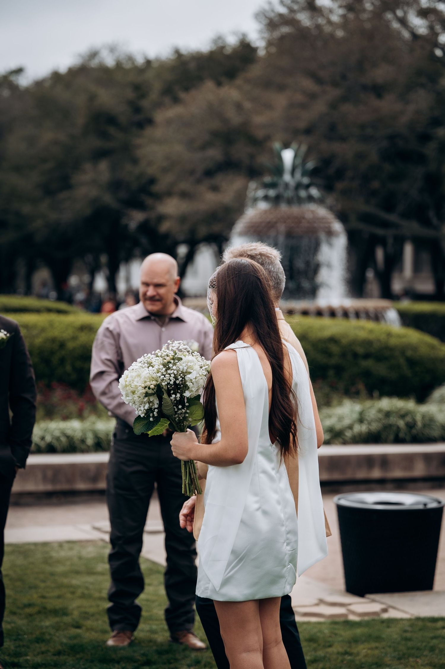 Bride in white mini dress holds white bouquet near fountain at outdoor wedding ceremony.