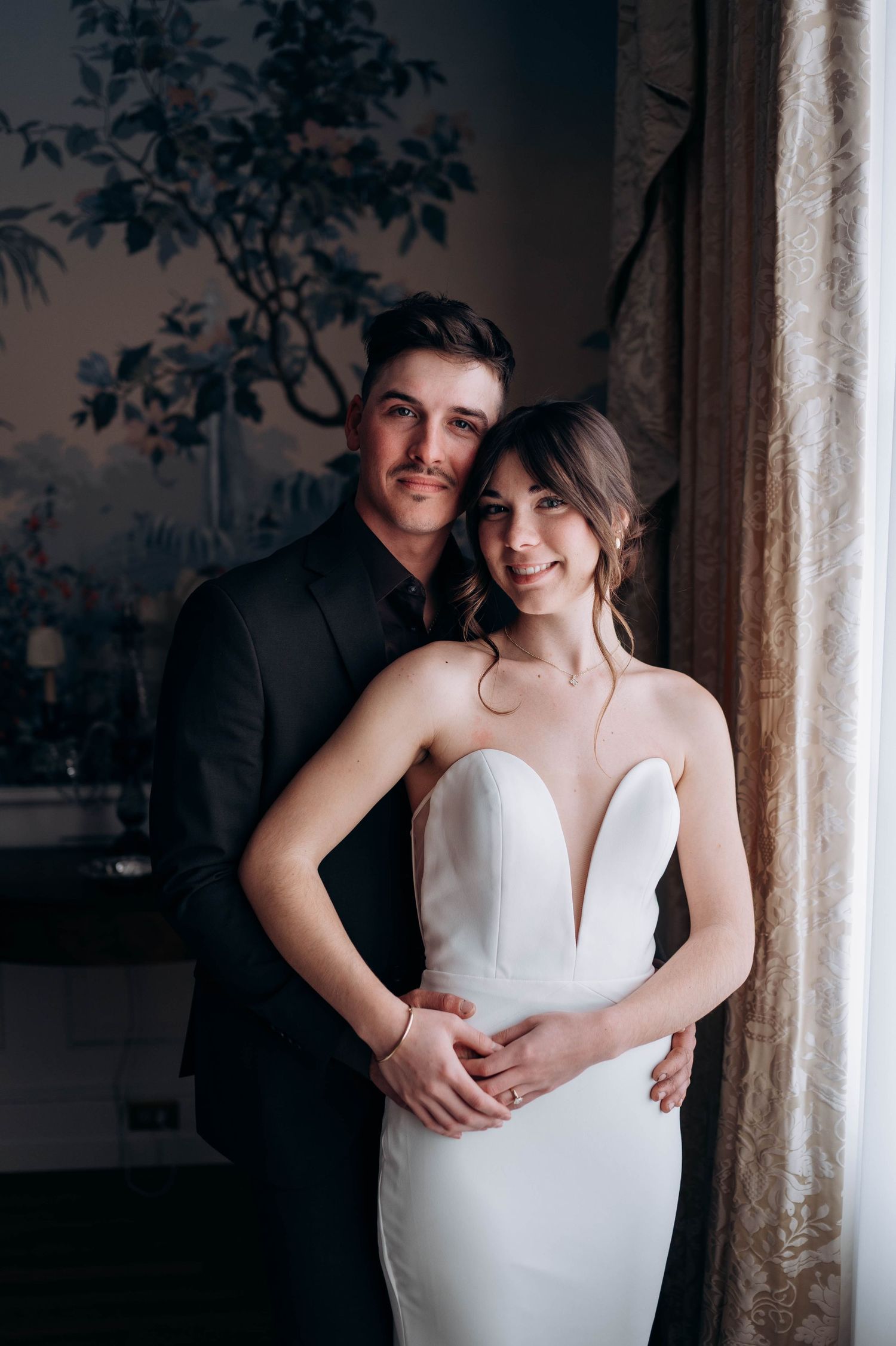 Bride and groom posing together in front of an ornate floral mural near a window.