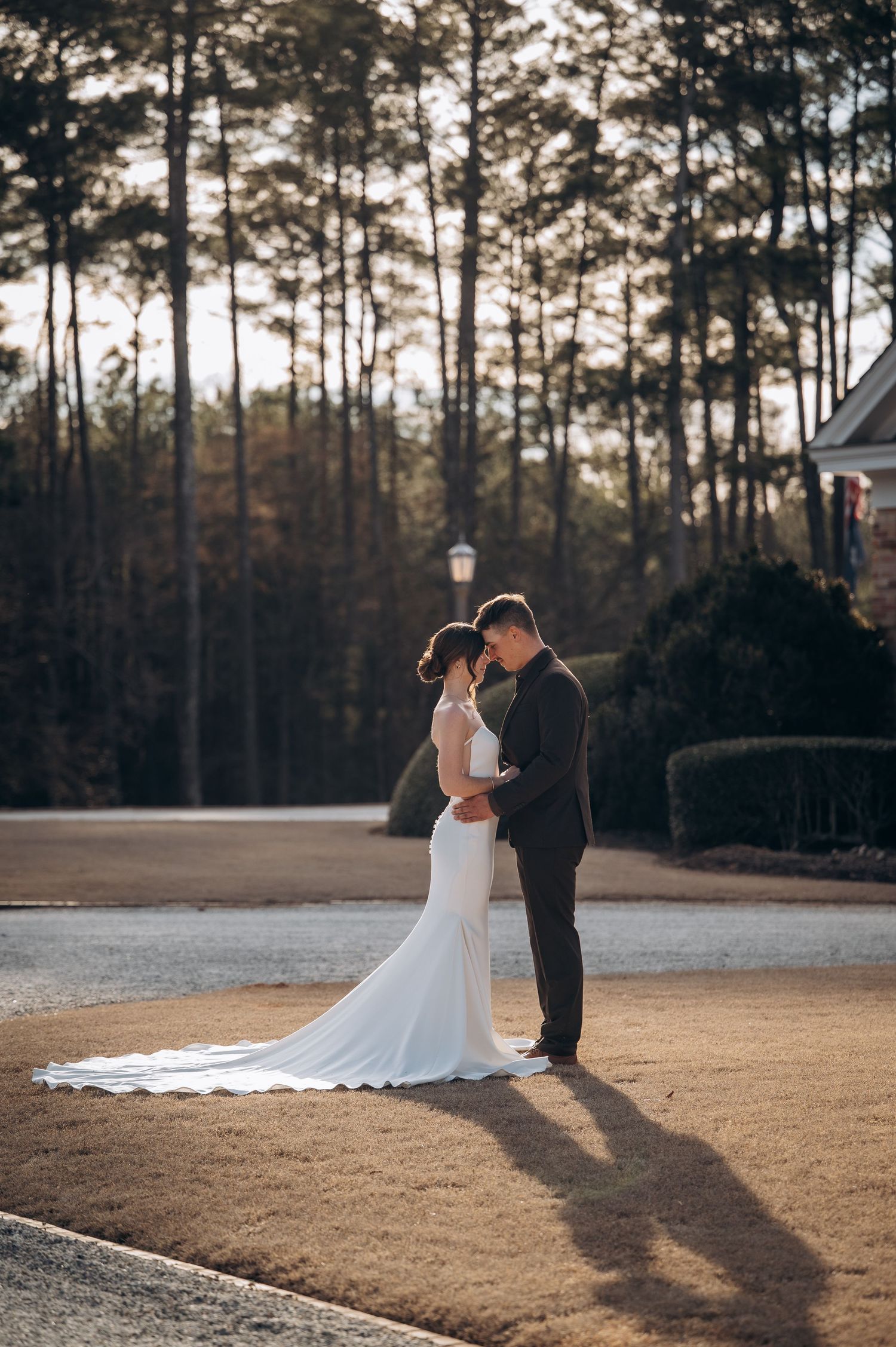 Bride and groom embrace at golden hour on a lawn surrounded by tall pine trees at an outdoor venue.