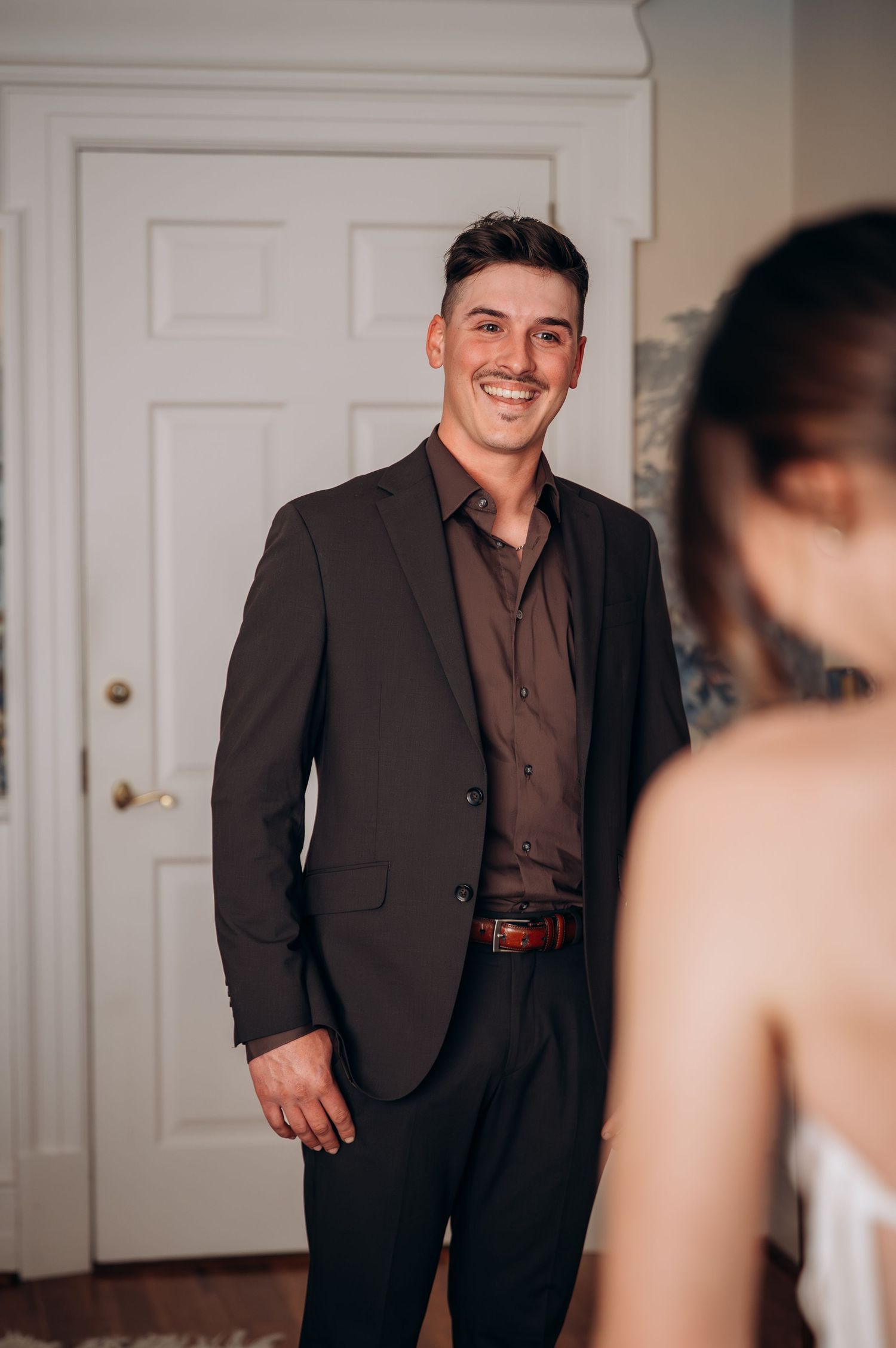 Smiling groom in dark suit reacts to seeing bride during first look moment indoors.