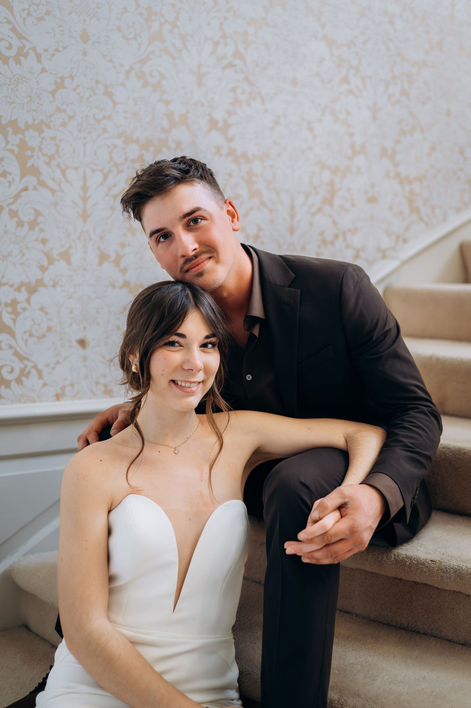Bride and groom sitting on elegant staircase, smiling at camera in wedding attire.