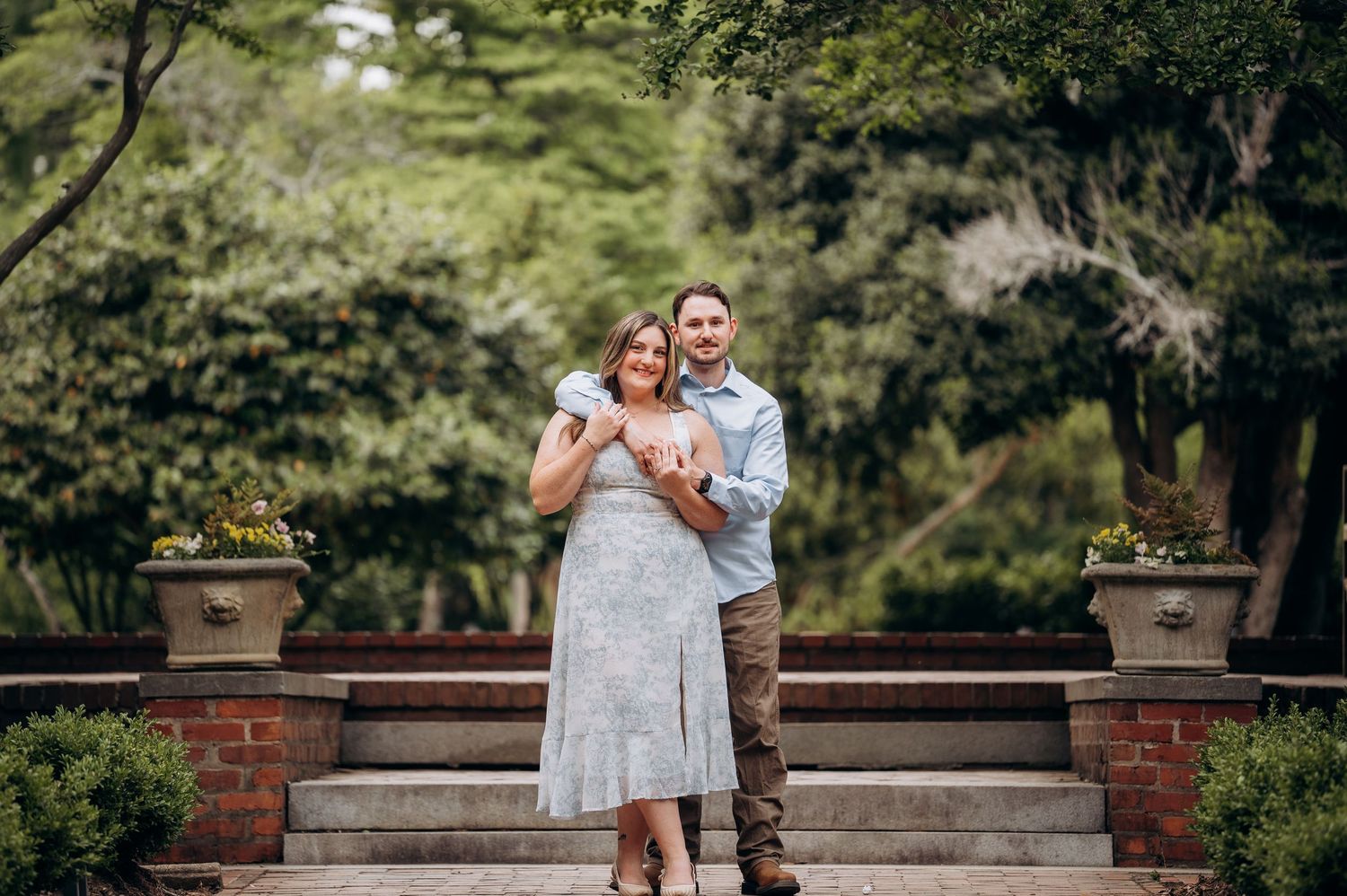 Couple embracing on garden steps with green trees and brick planters in a serene outdoor setting.