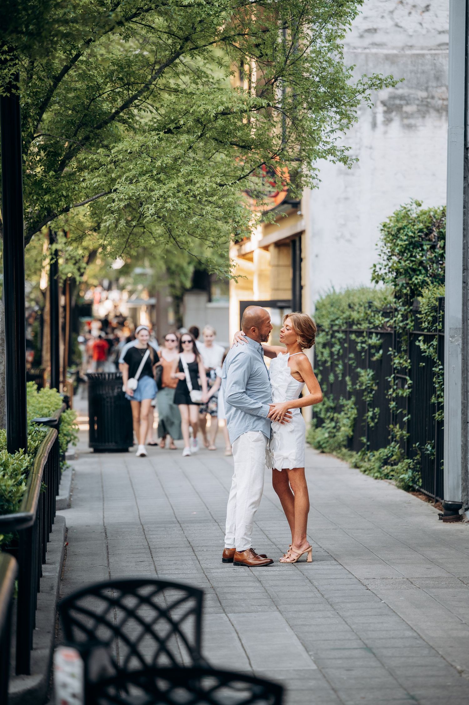 A couple embraces on a tree-lined urban sidewalk, dressed in white and light blue summer outfits.