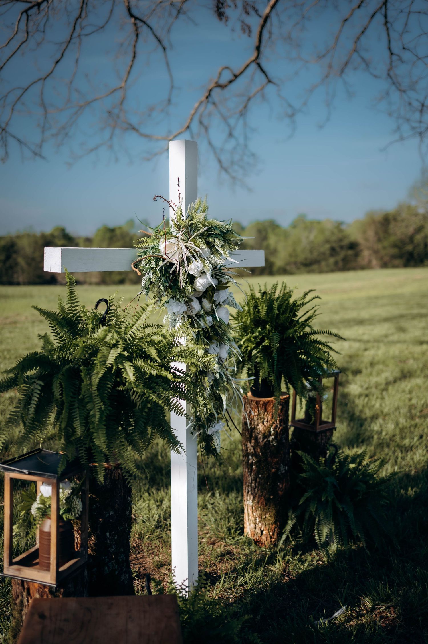 White cross adorned with ferns and greenery stands in an open field, surrounded by rustic wood stumps and a lantern.
