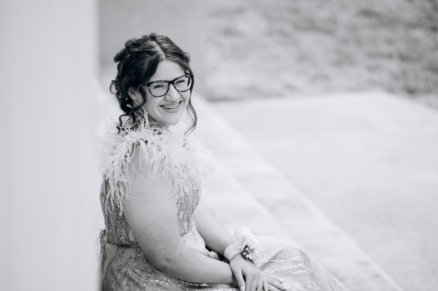 Black and white photo of a smiling bride in a feathered dress and glasses, seated outdoors on her wedding day.