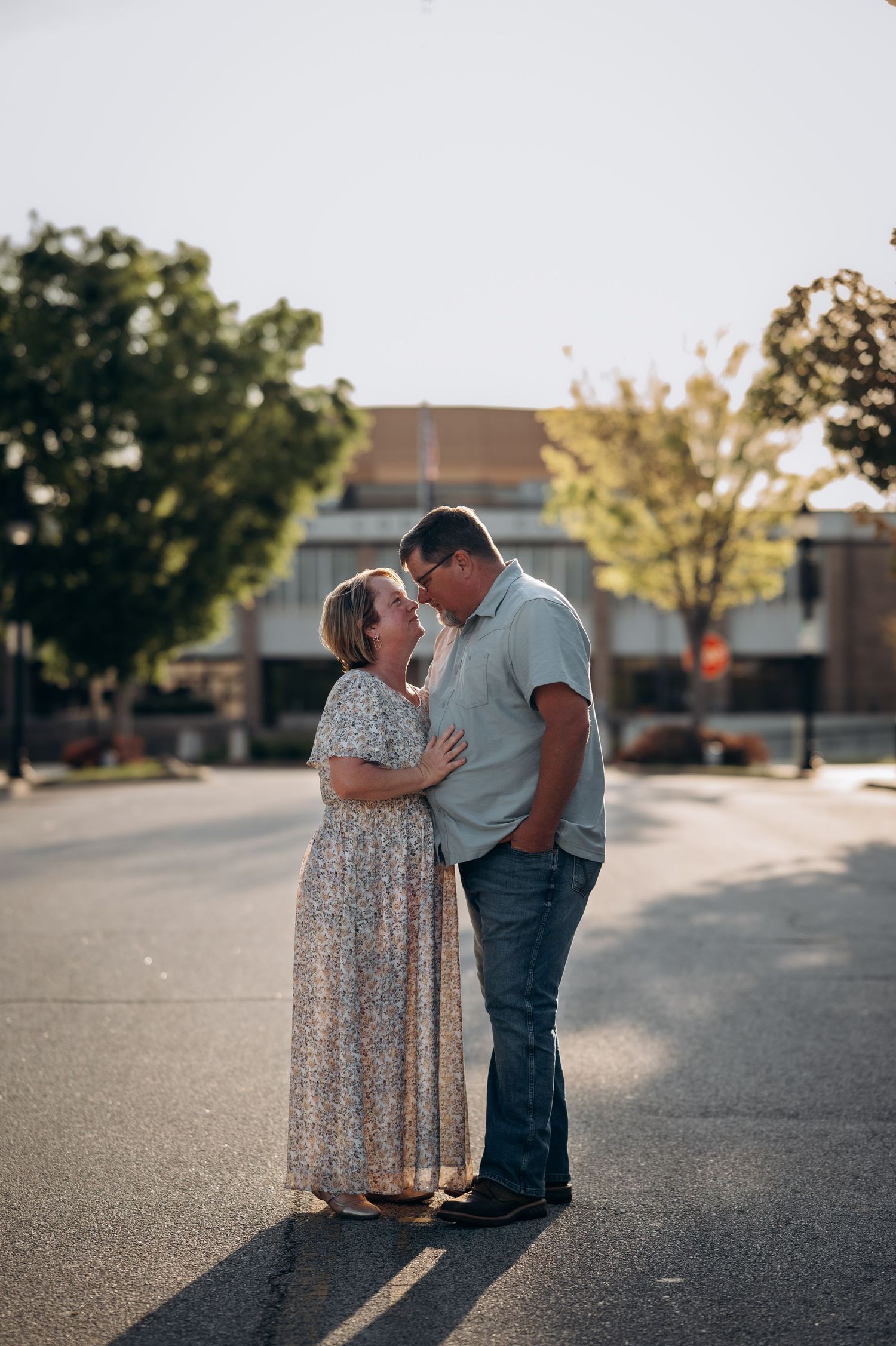 Couple sharing a romantic moment kissing in a sunny street, woman in floral maxi dress, man in blue shirt and jeans.