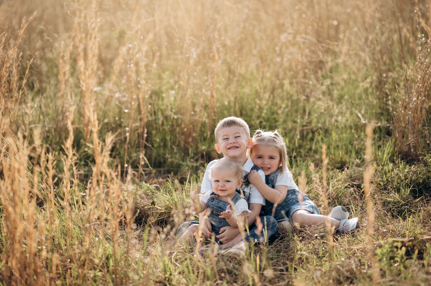 Three young children sit together in a sunny golden field, smiling warmly in a heartwarming outdoor portrait.