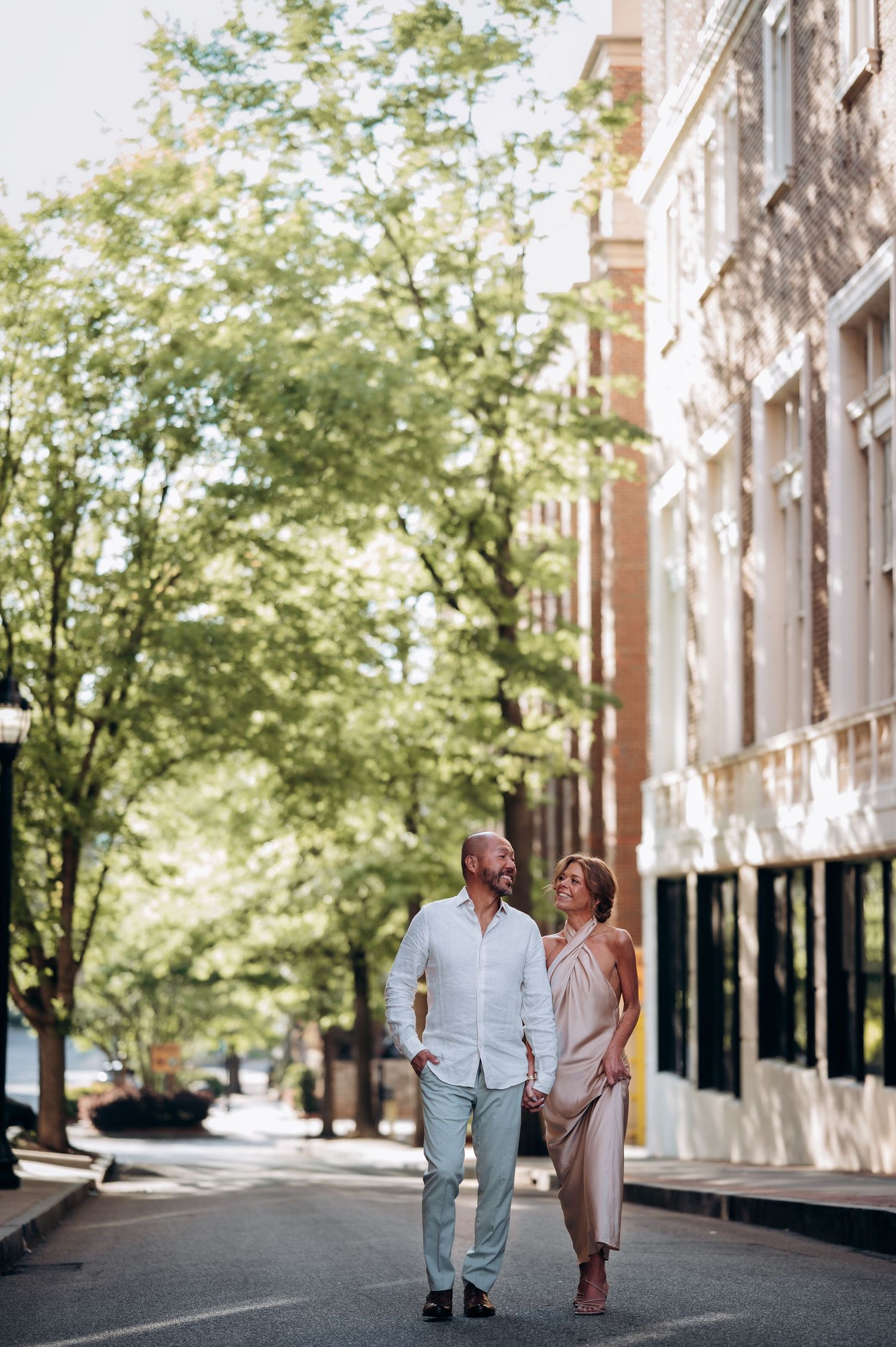 Well-dressed couple walking hand-in-hand down a sunny urban street lined with green trees and brick buildings.