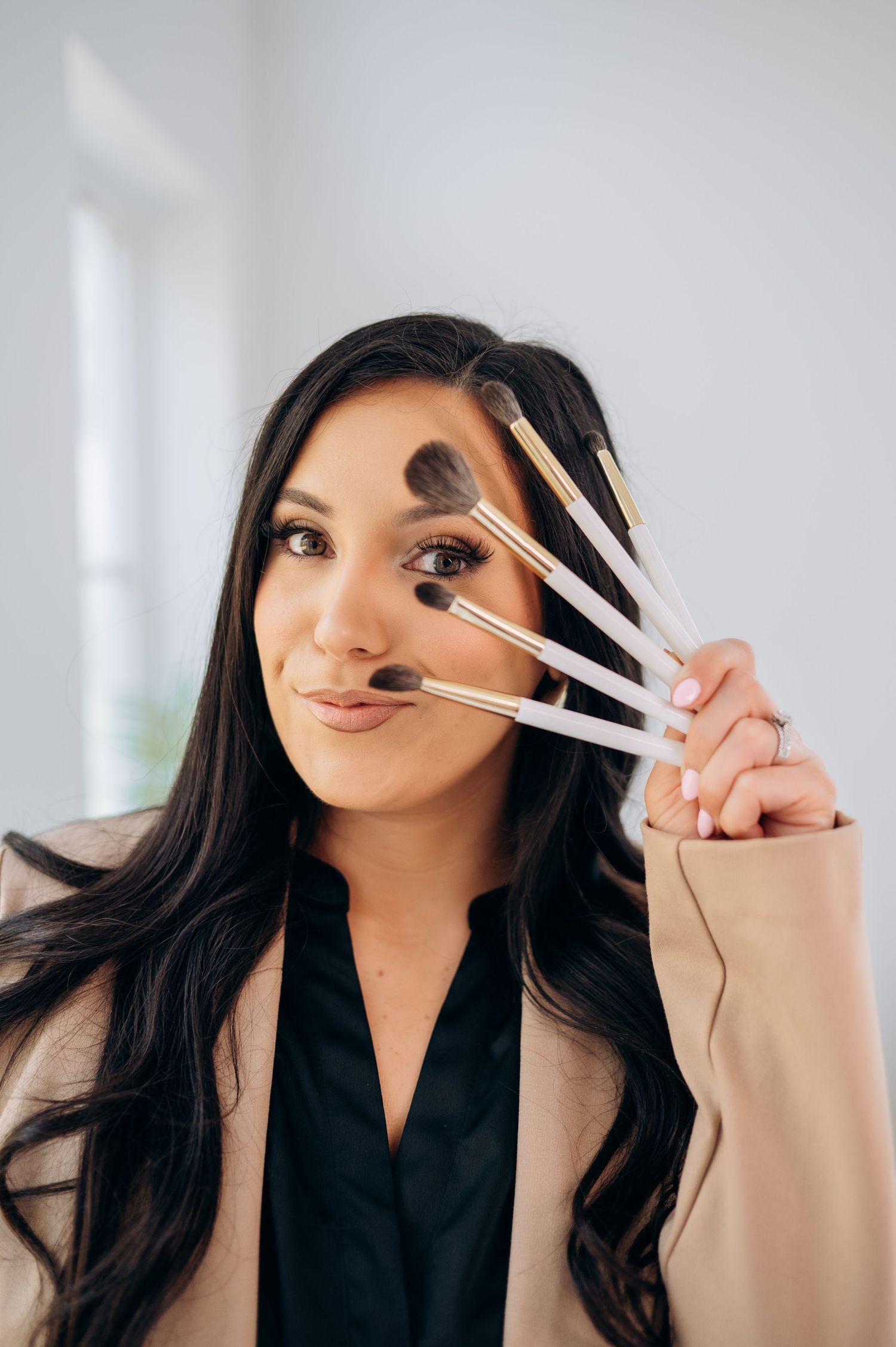 Playful beauty portrait holding multiple makeup brushes fanned out against white wall.