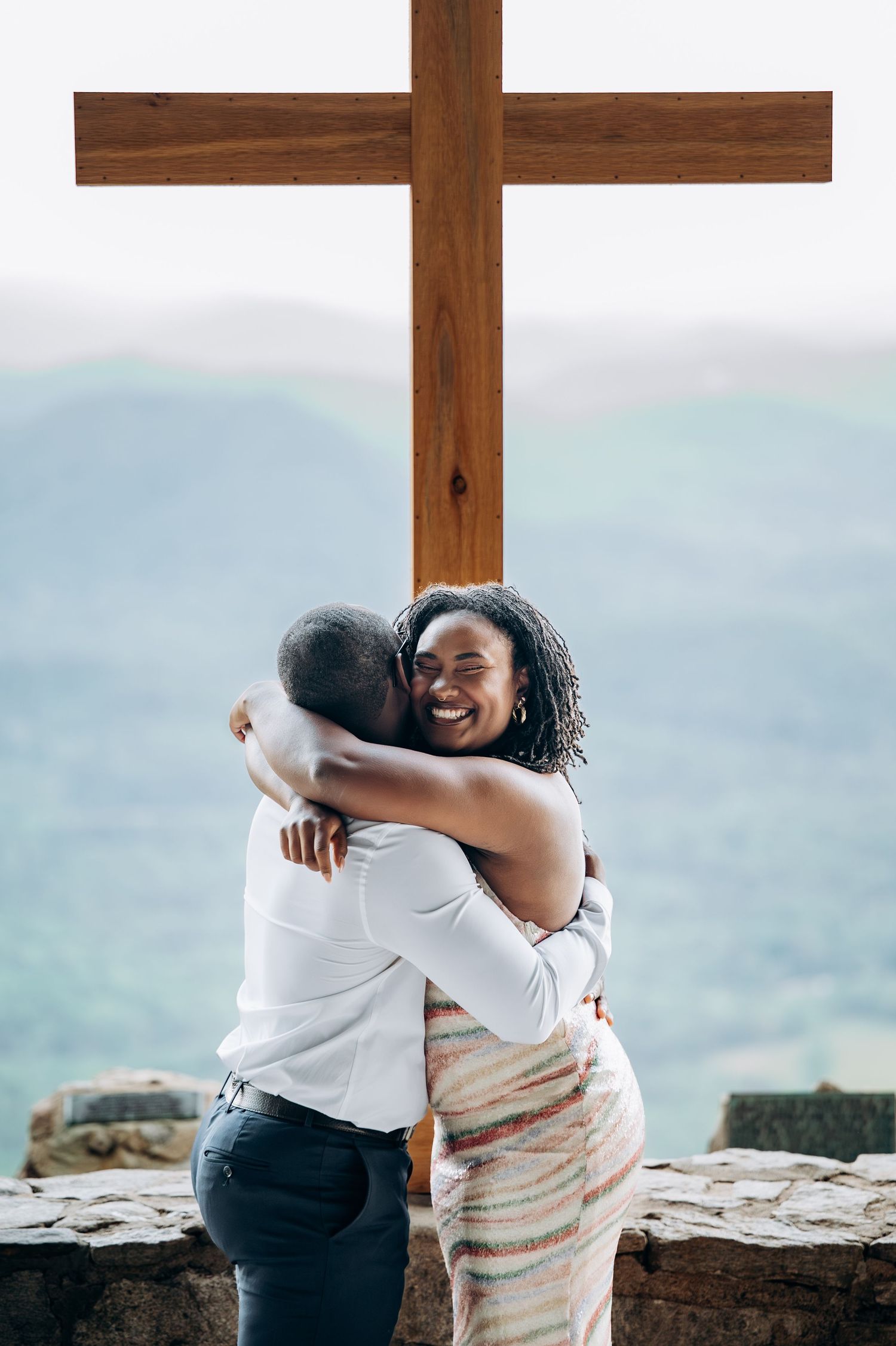 Couple embracing and smiling in front of a wooden cross with scenic mountain views in the background.