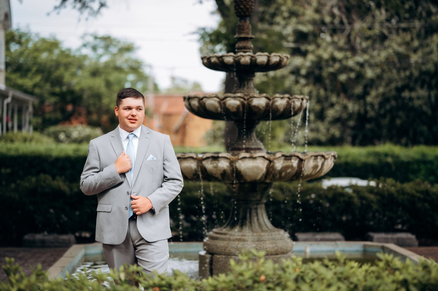 Man in gray suit posing beside ornate stone tiered fountain in lush garden setting.