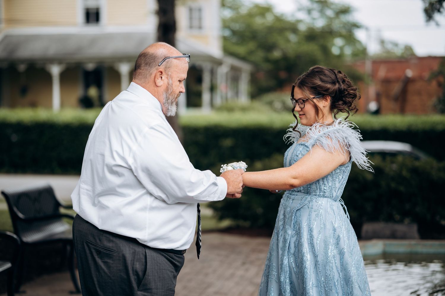 A man and woman in formal attire dance outdoors near a fountain, she wearing a blue feathered dress.