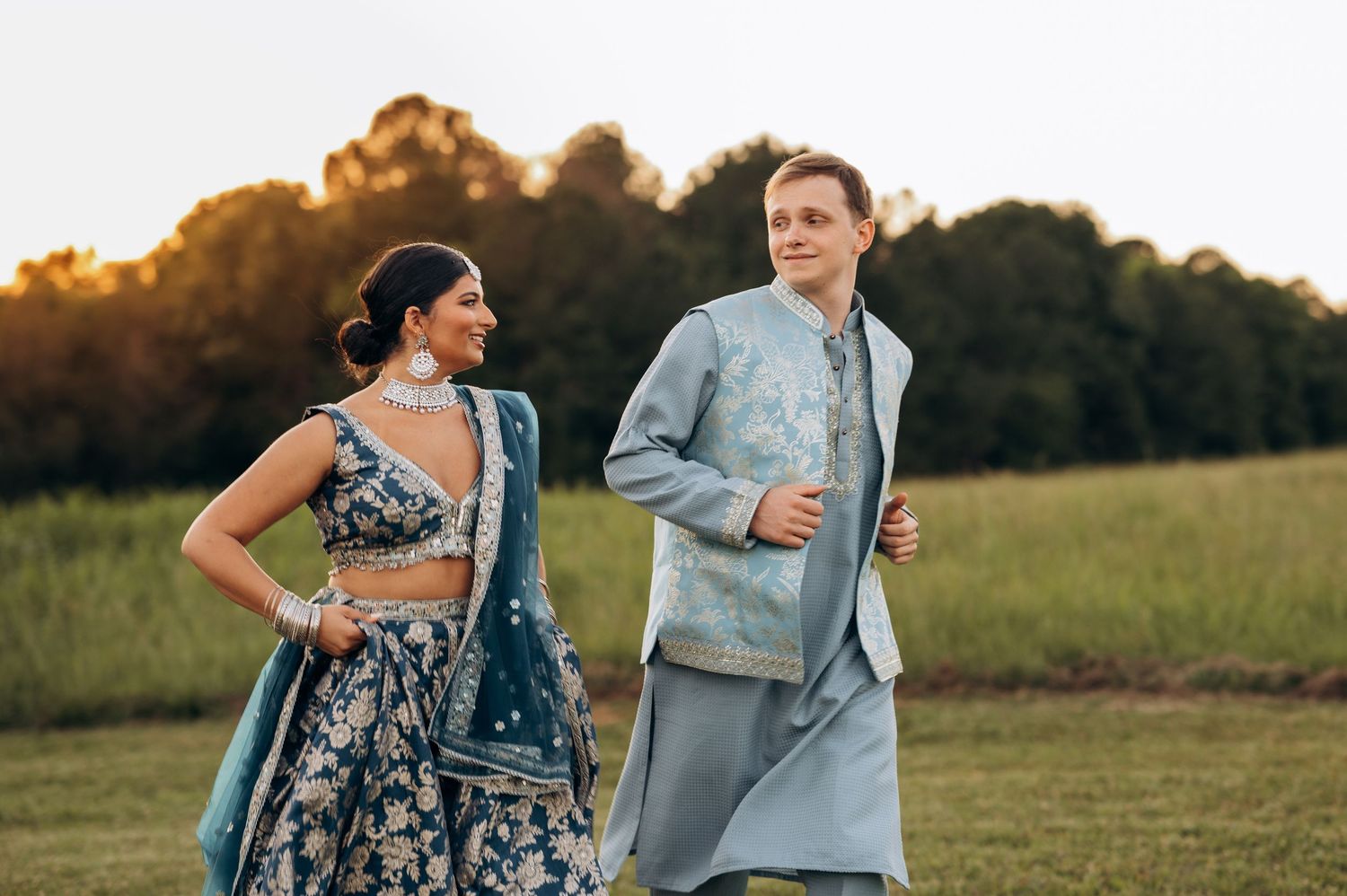 Couple in traditional Indian wedding attire walking outdoors at sunset, woman in blue lehenga, man in matching sherwani.