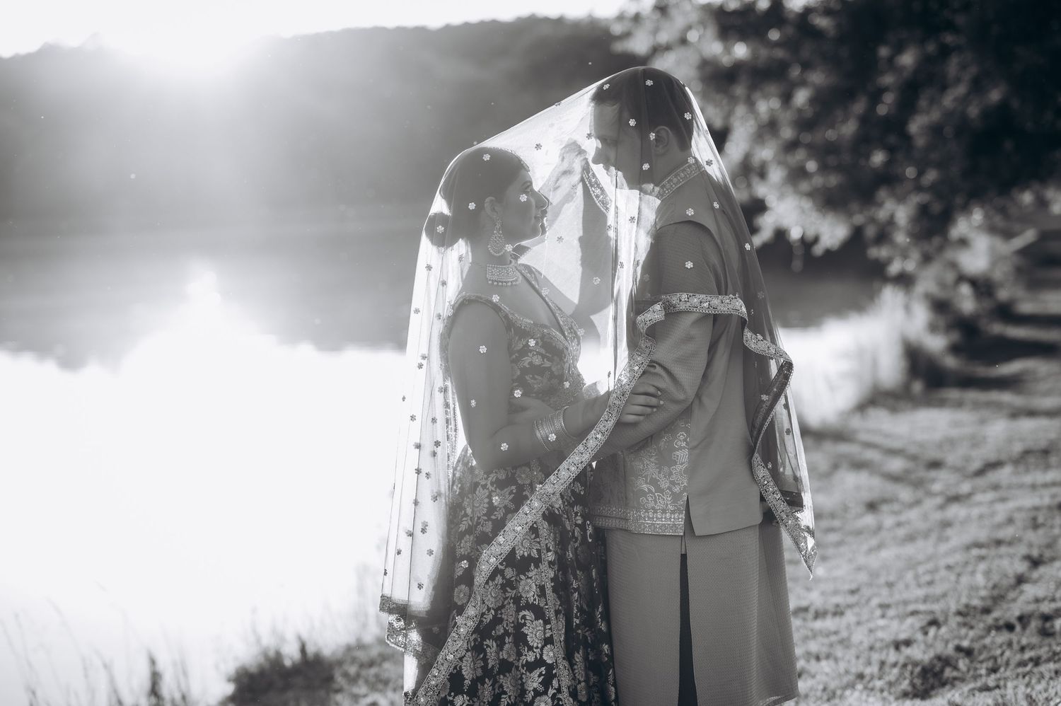 Black and white photo of Indian bride and groom sharing intimate moment under veil by lakeside at golden hour.