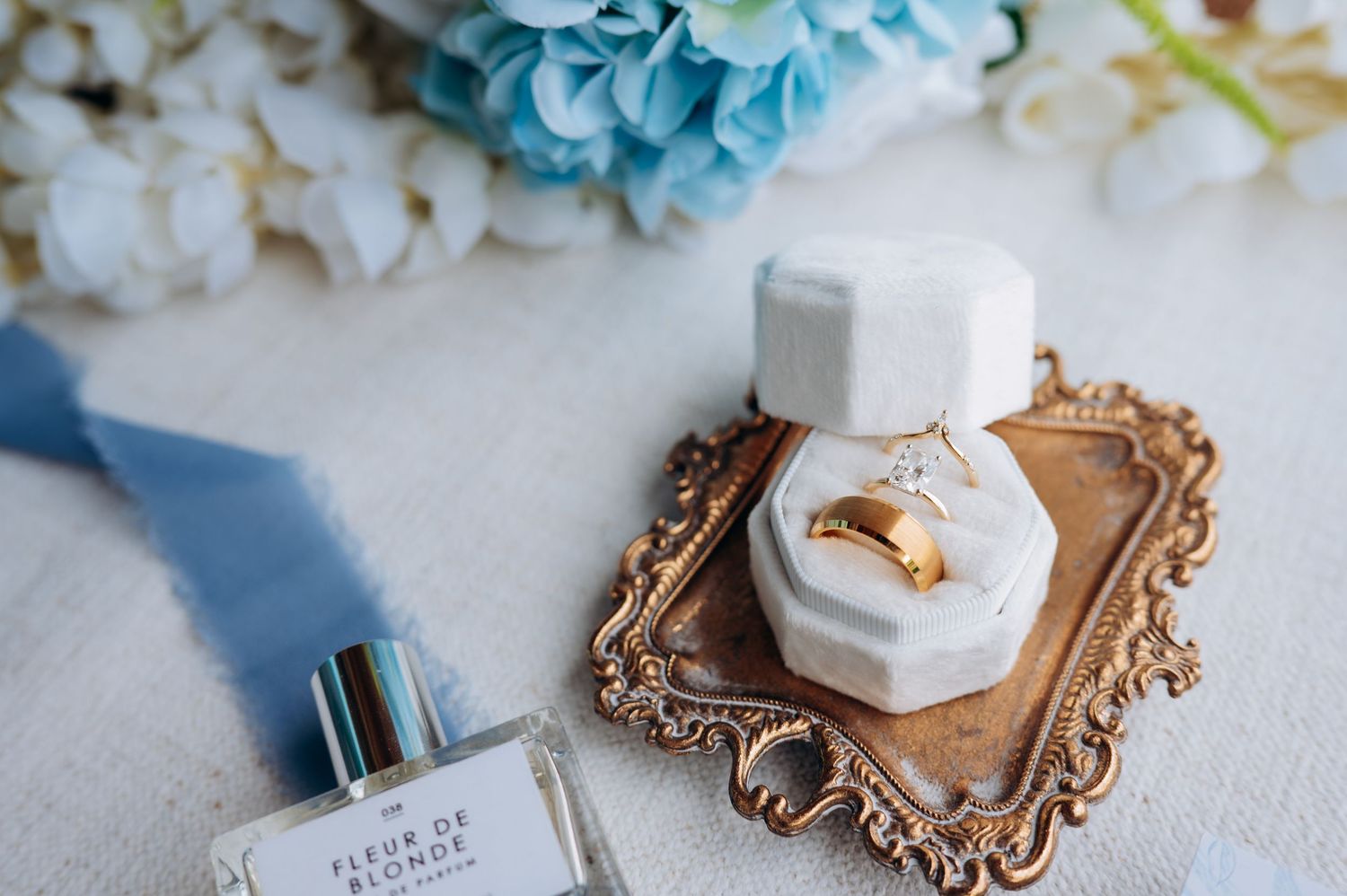 Wedding flat lay with gold ring in velvet box, Fleur de Blondie perfume, and blue floral accents.
