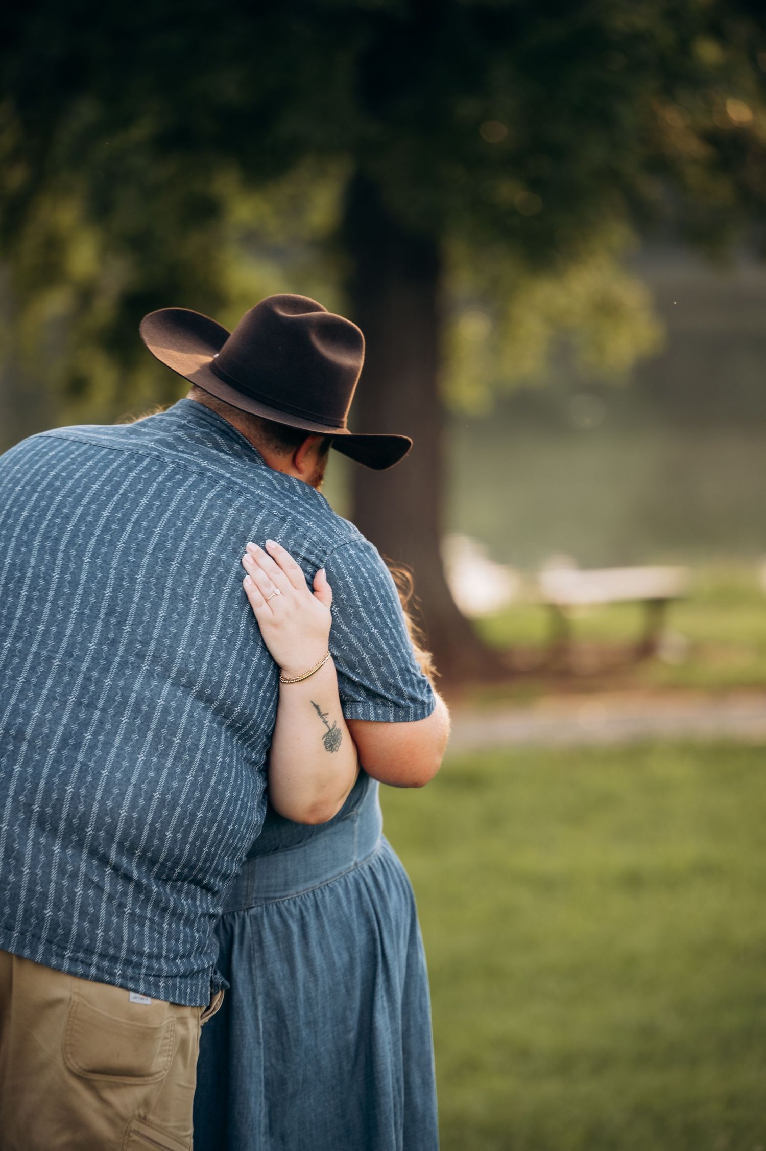 A man in a brown cowboy hat and blue shirt embraces a woman in a gray dress outdoors near a serene park.