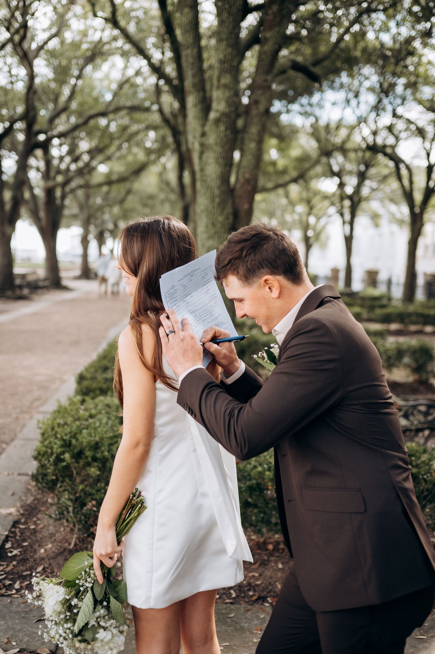 Groom signing marriage license using bride's back as a writing surface in a tree-lined park.
