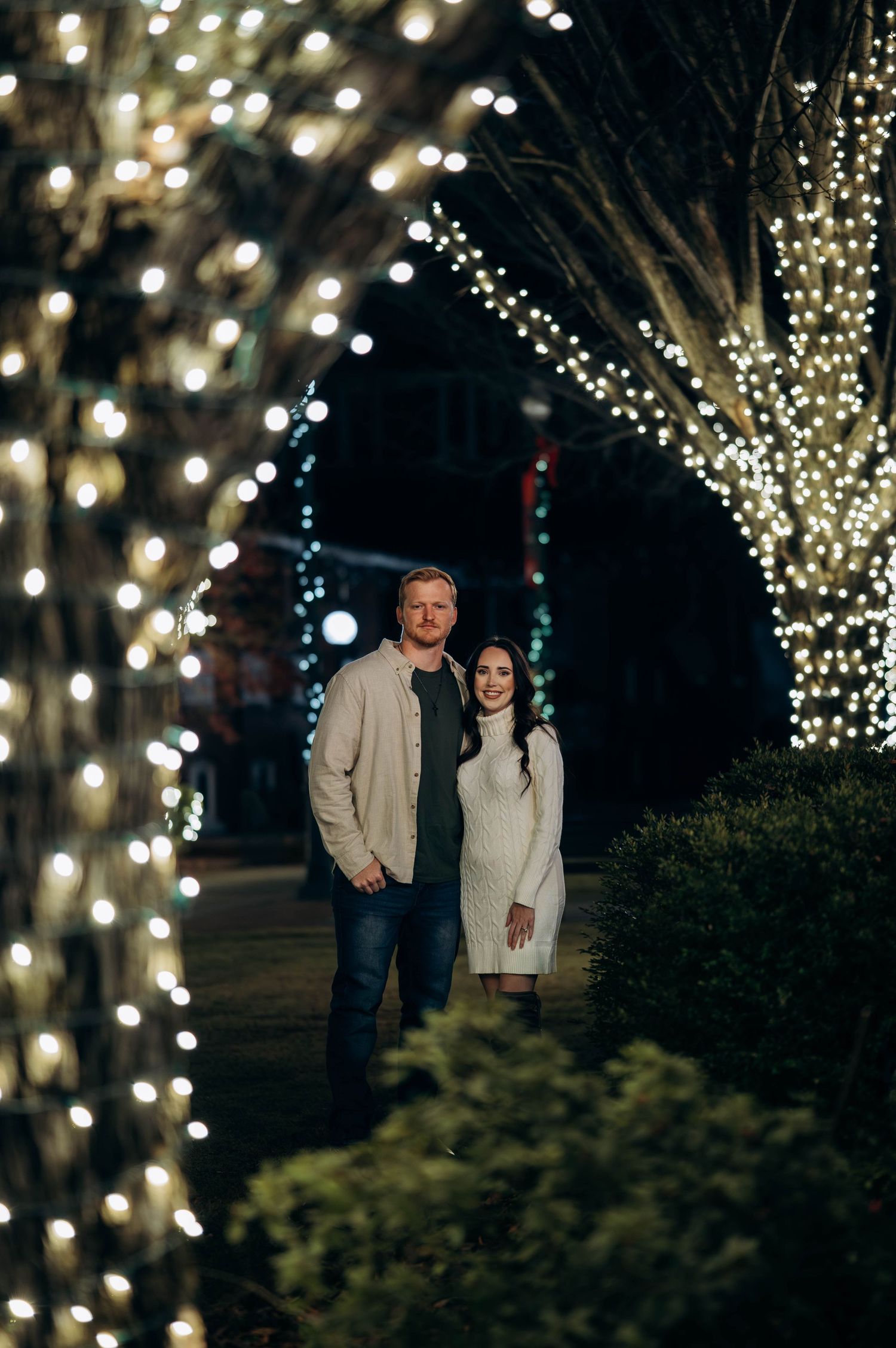Couple standing among trees wrapped in white fairy lights at night, woman in white dress and man in beige jacket.