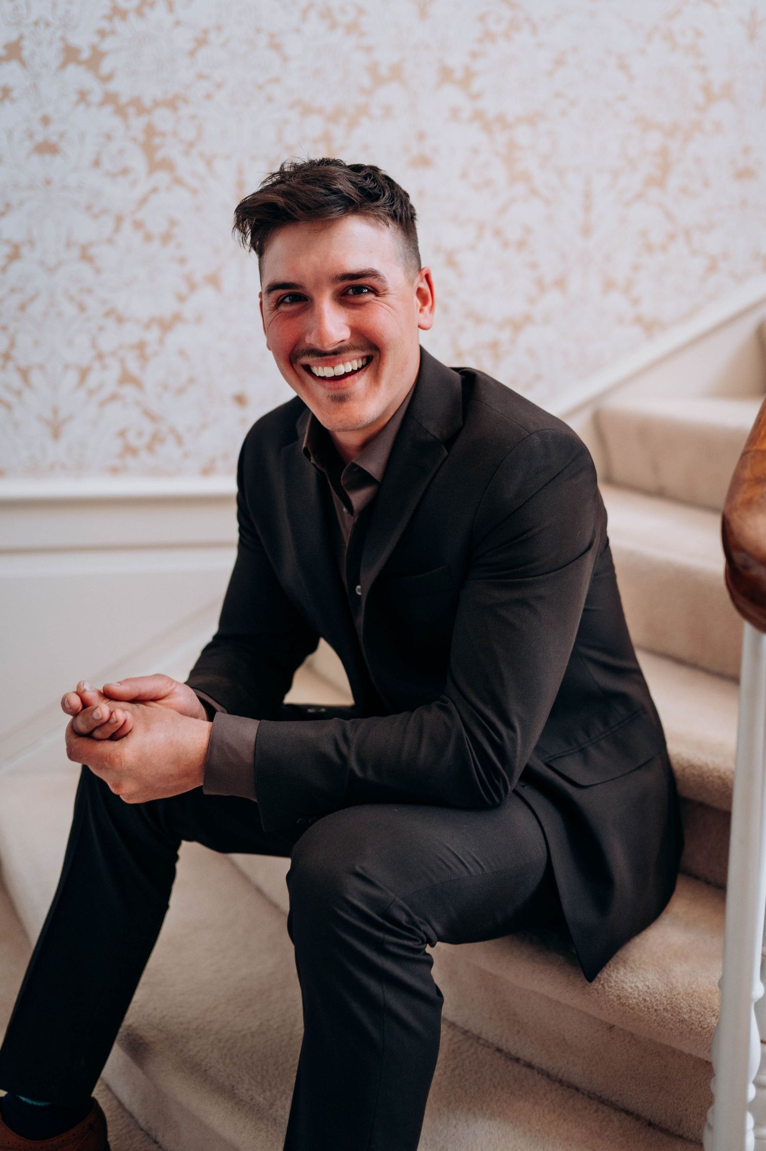 Smiling groom in dark suit sits on carpeted stairs against elegant gold damask wallpaper.