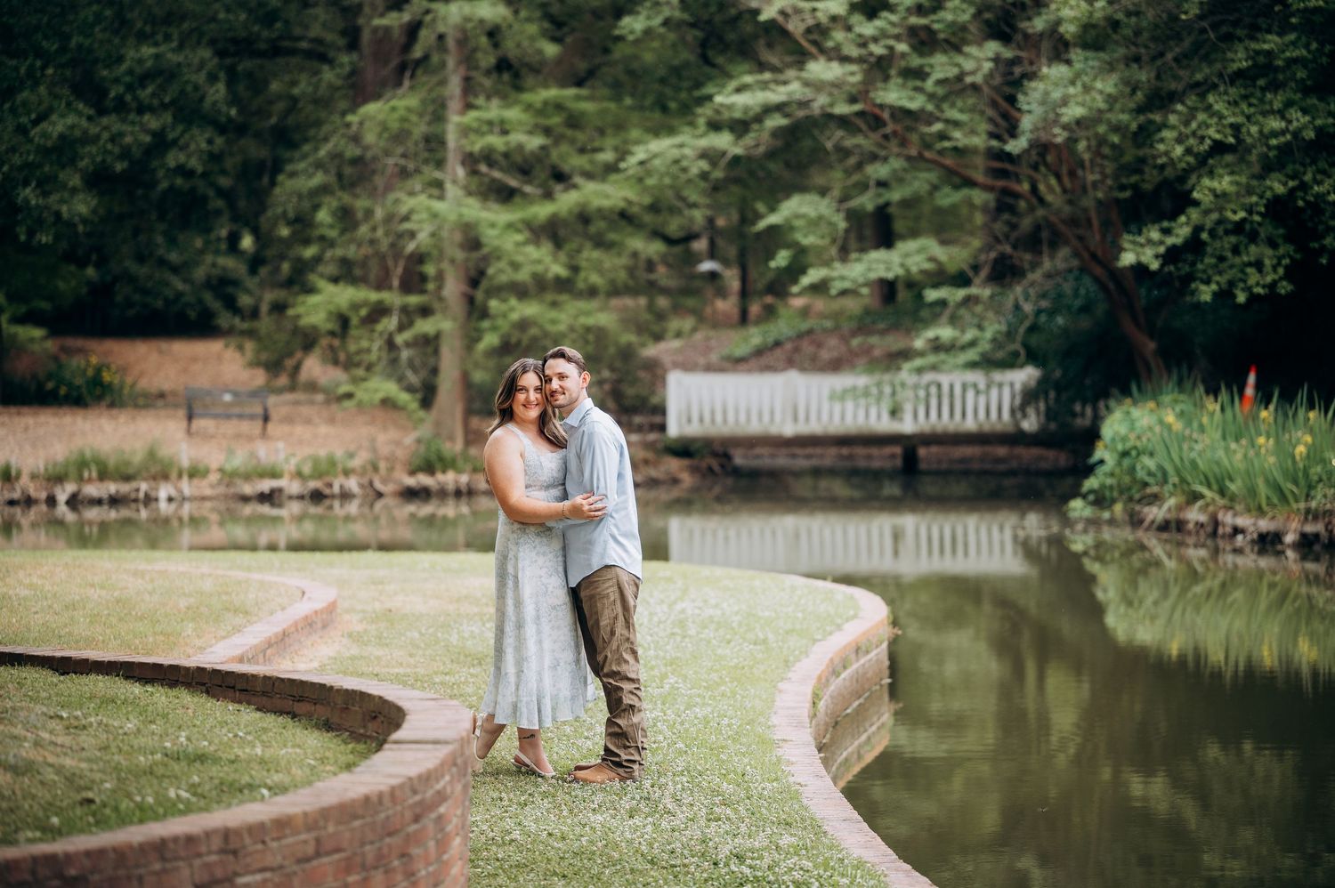 Happy couple embracing near a curved garden pond with lush green trees in background.