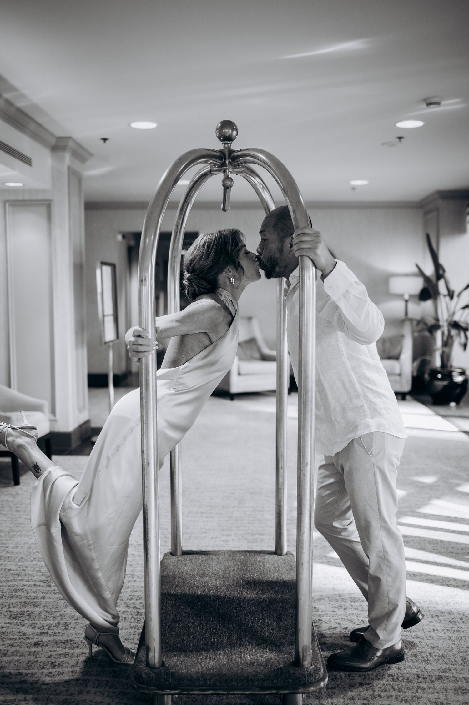 Black and white photo of couple kissing through a hotel luggage cart.