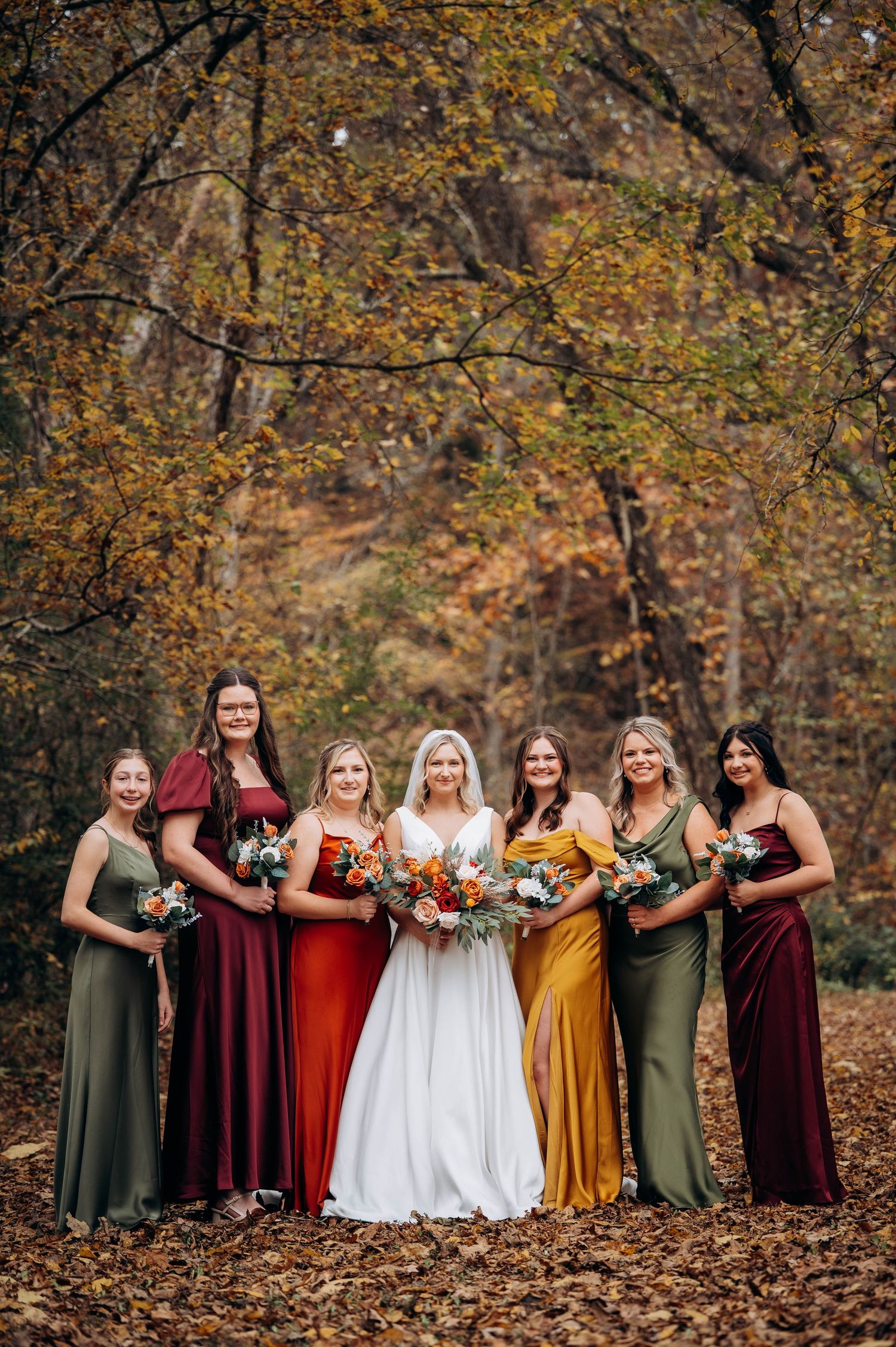 Bridal party in jewel-toned dresses poses among autumn foliage.
