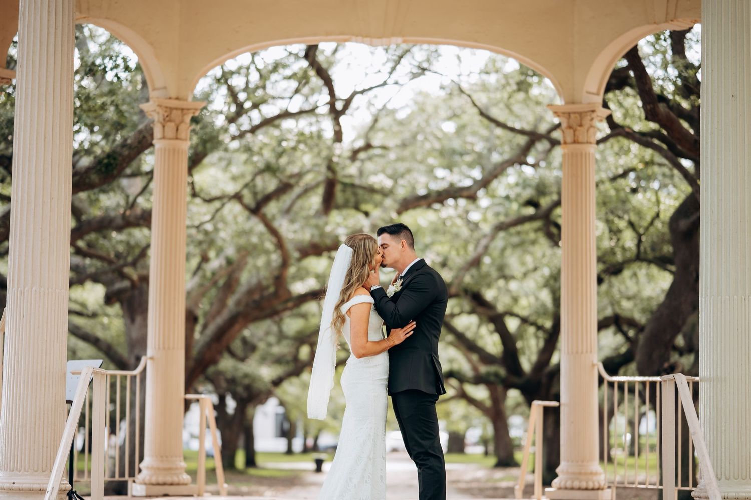 Bride and groom share a romantic embrace under elegant columns surrounded by lush oak trees at an outdoor wedding venue.