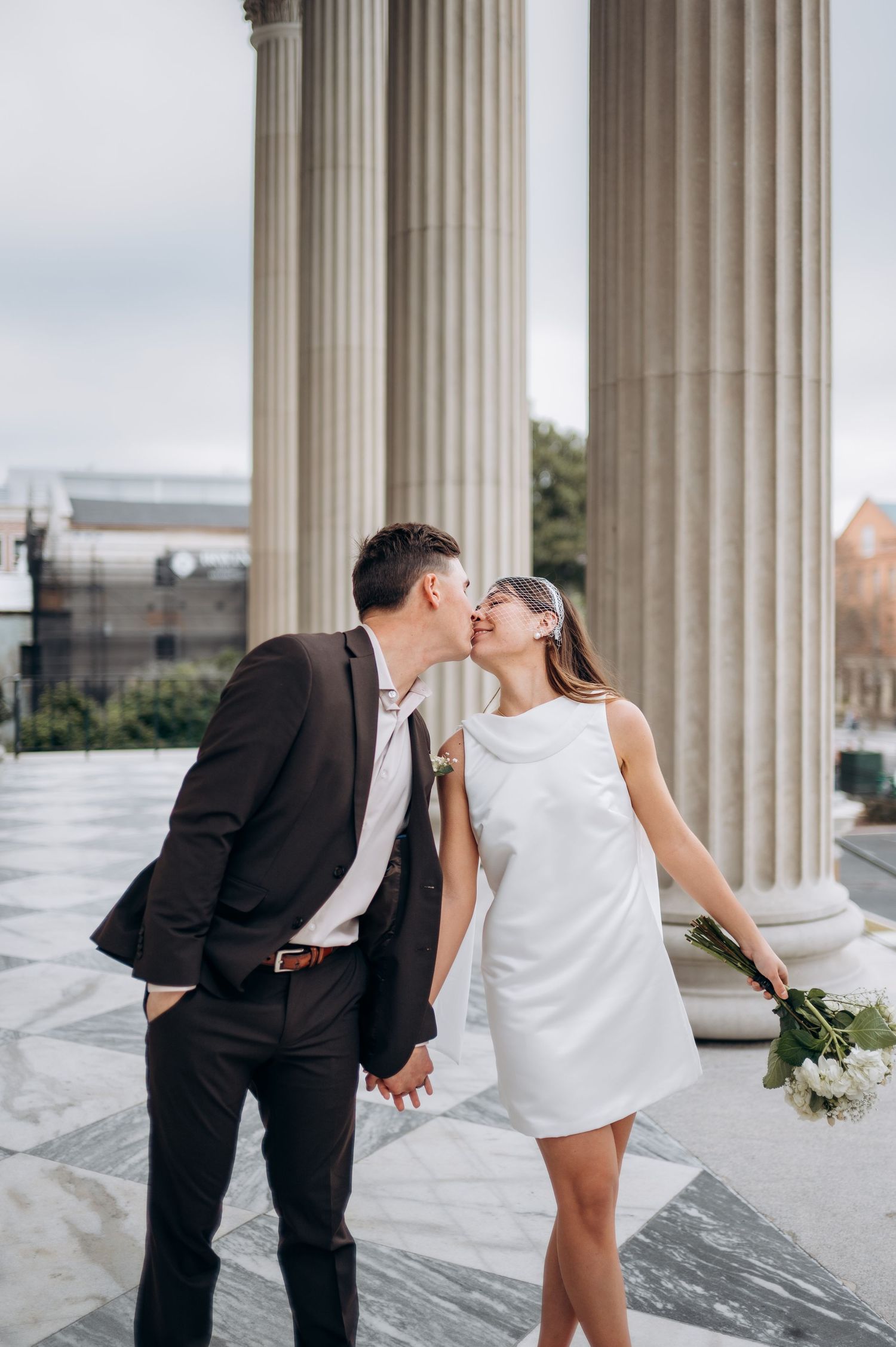 A romantic couple shares playful kisses between classic white marble columns in an elegant architectural setting.
