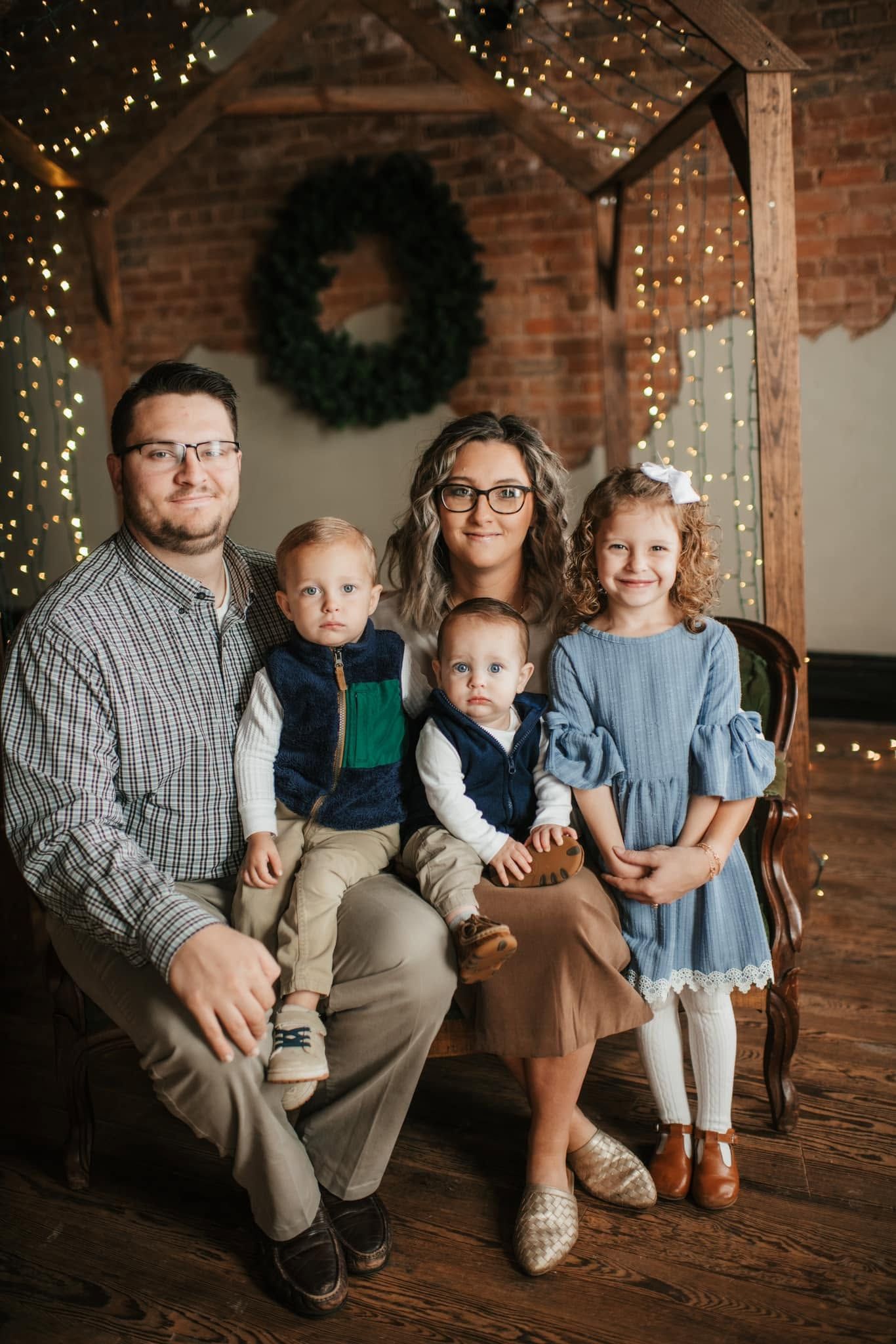 Family of five posing for a Christmas portrait with festive lights and wreath backdrop.