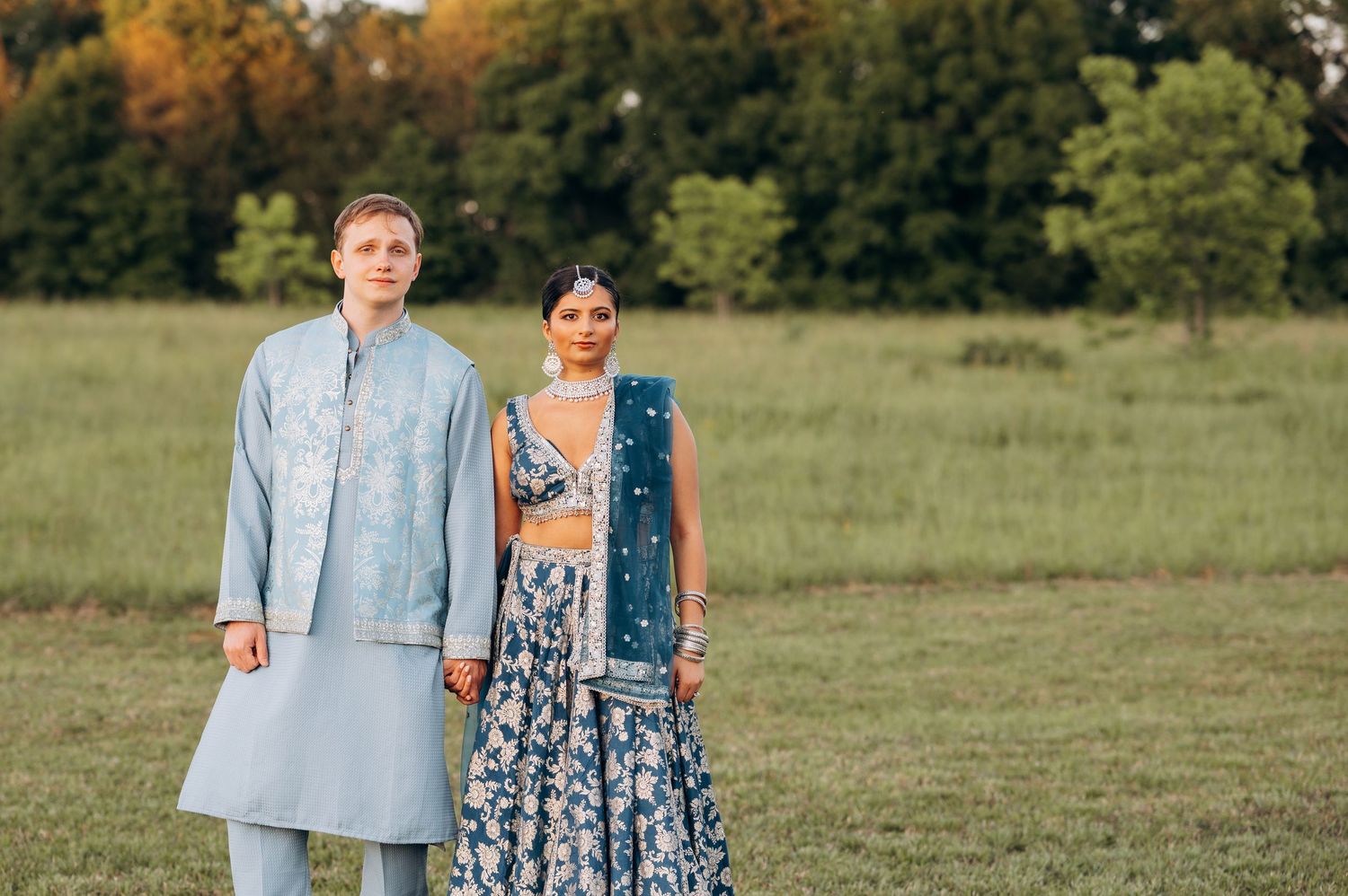 Couple in traditional South Asian wedding attire standing in a green field, man in light blue kurta, woman in blue lehenga.