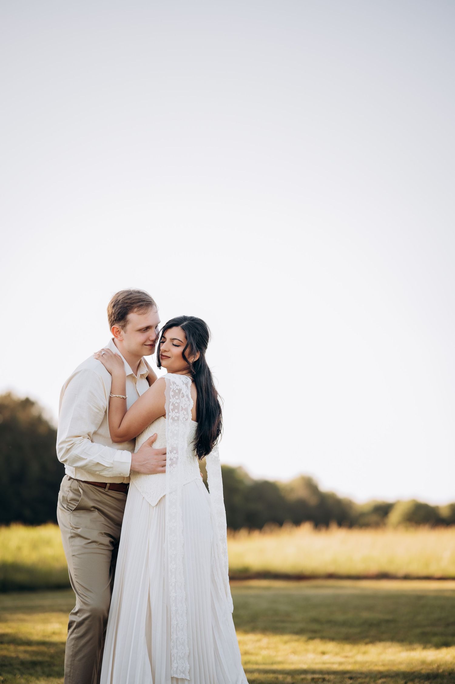 Bride in white lace gown embraces groom in beige suit during golden hour outdoor wedding portrait.