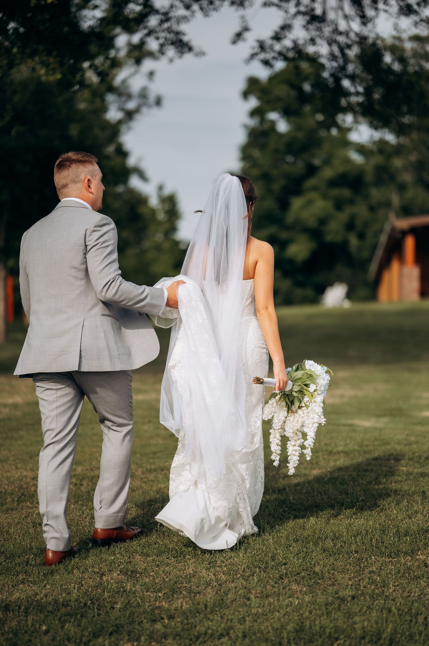 Bride and groom walking hand in hand on green grass, bride wearing white gown with veil and floral bouquet.