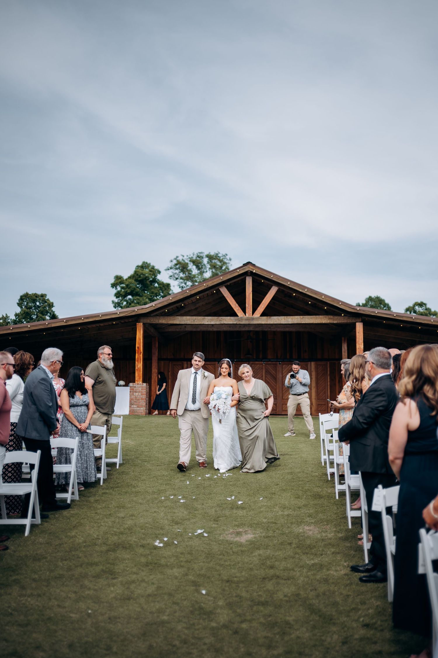 Outdoor wedding ceremony on green lawn with rustic barn backdrop and guests lining the aisle.