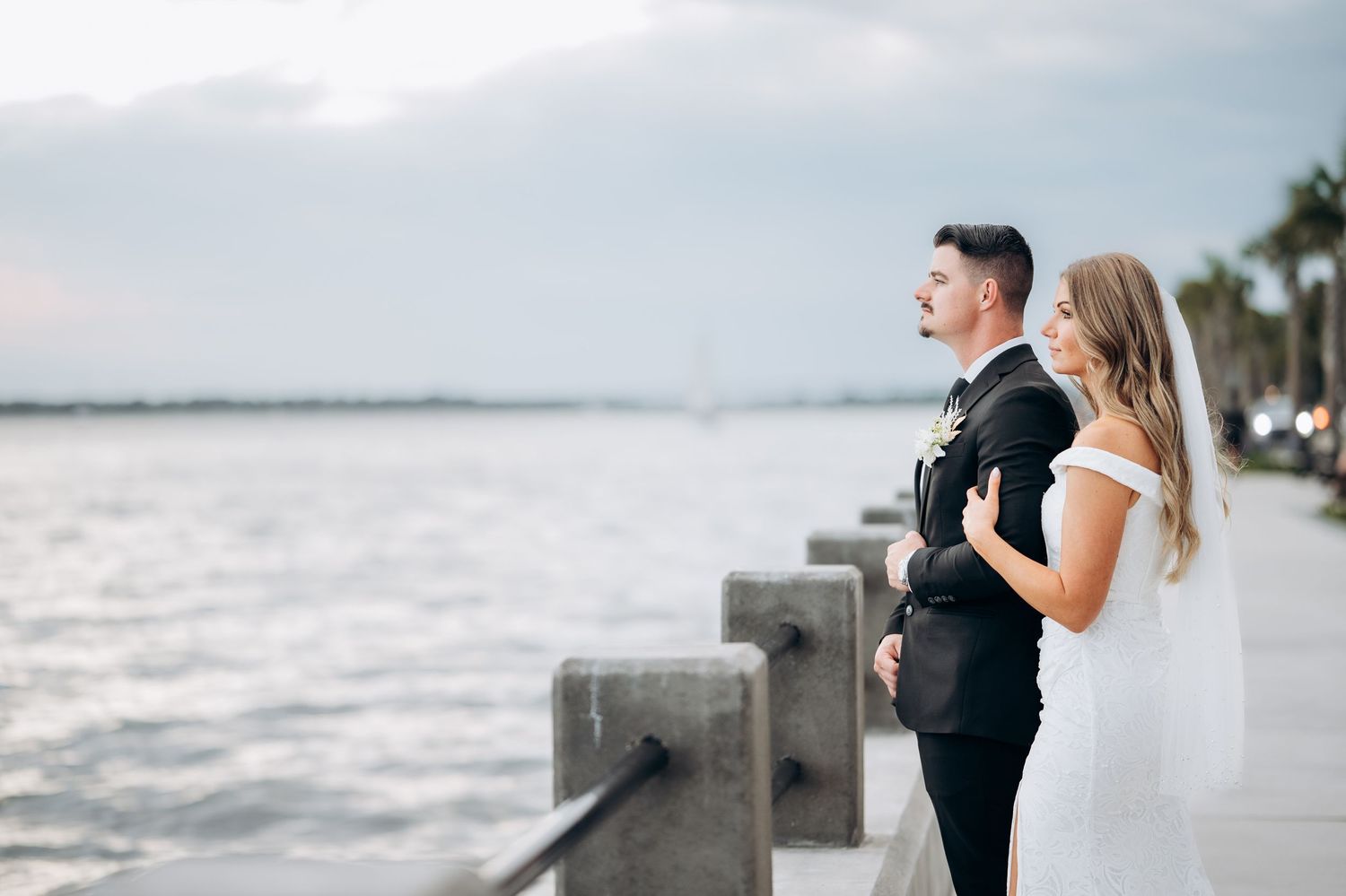 Bride and groom standing together on a waterfront promenade at dusk, gazing at the scenic ocean view.