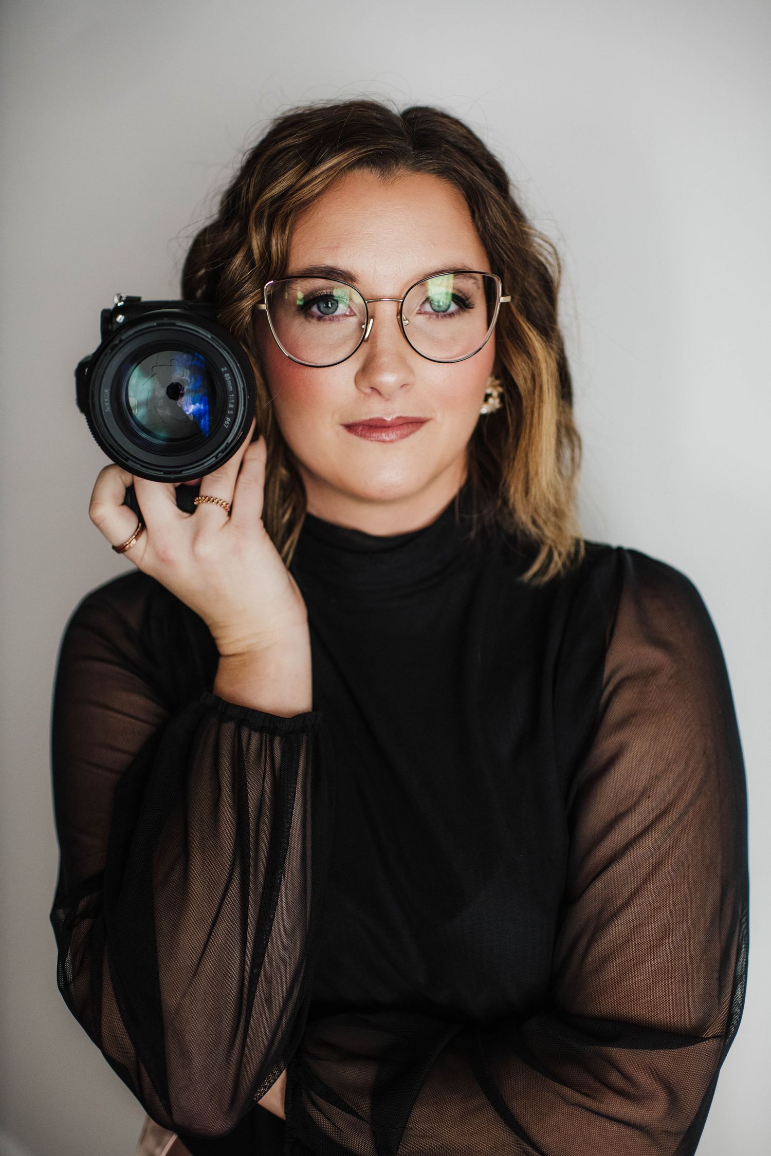 Photographer in black sheer top and glasses poses with camera against white backdrop.