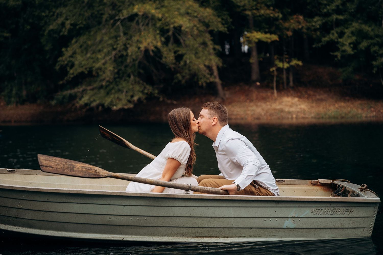 A romantic couple shares intimate moments in a rowboat on a tranquil lake surrounded by trees.